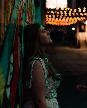A moody nighttime portrait with string lights bokeh, illuminating a woman in a white dress leaning against a turquoise wall.