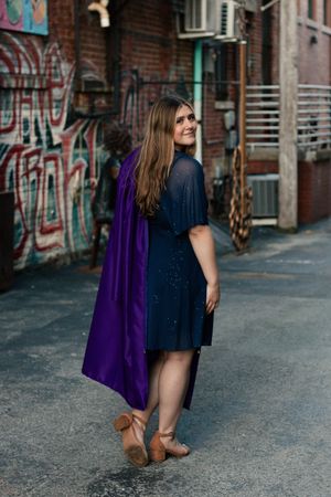 A person in a blue dress and purple cape stands in an urban alley with graffiti walls and brown boots.