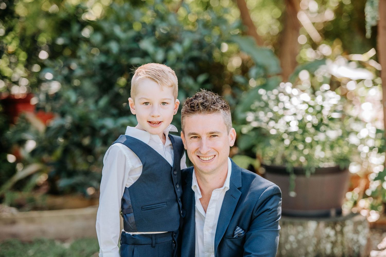 A formal outdoor portrait shows two people in suits standing together in a garden setting.