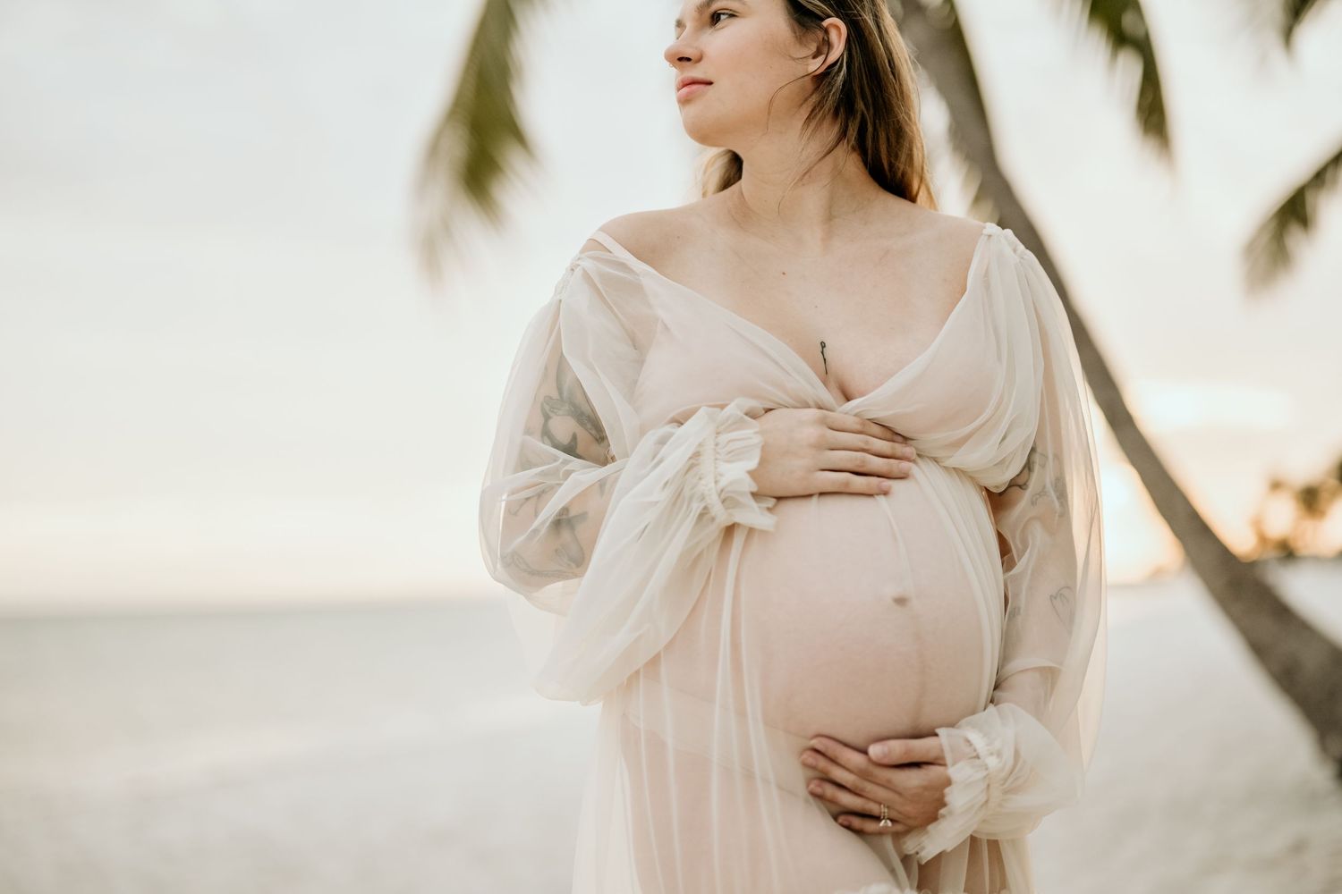 Dreamy beach maternity photo in flowing white dress with palm trees at sunset.