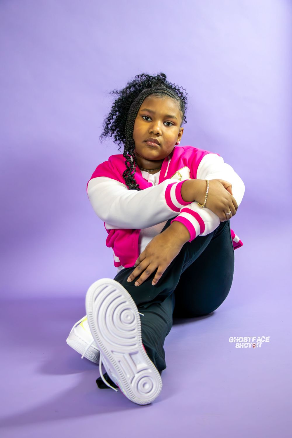 Young person in pink varsity jacket and black pants poses against purple backdrop.