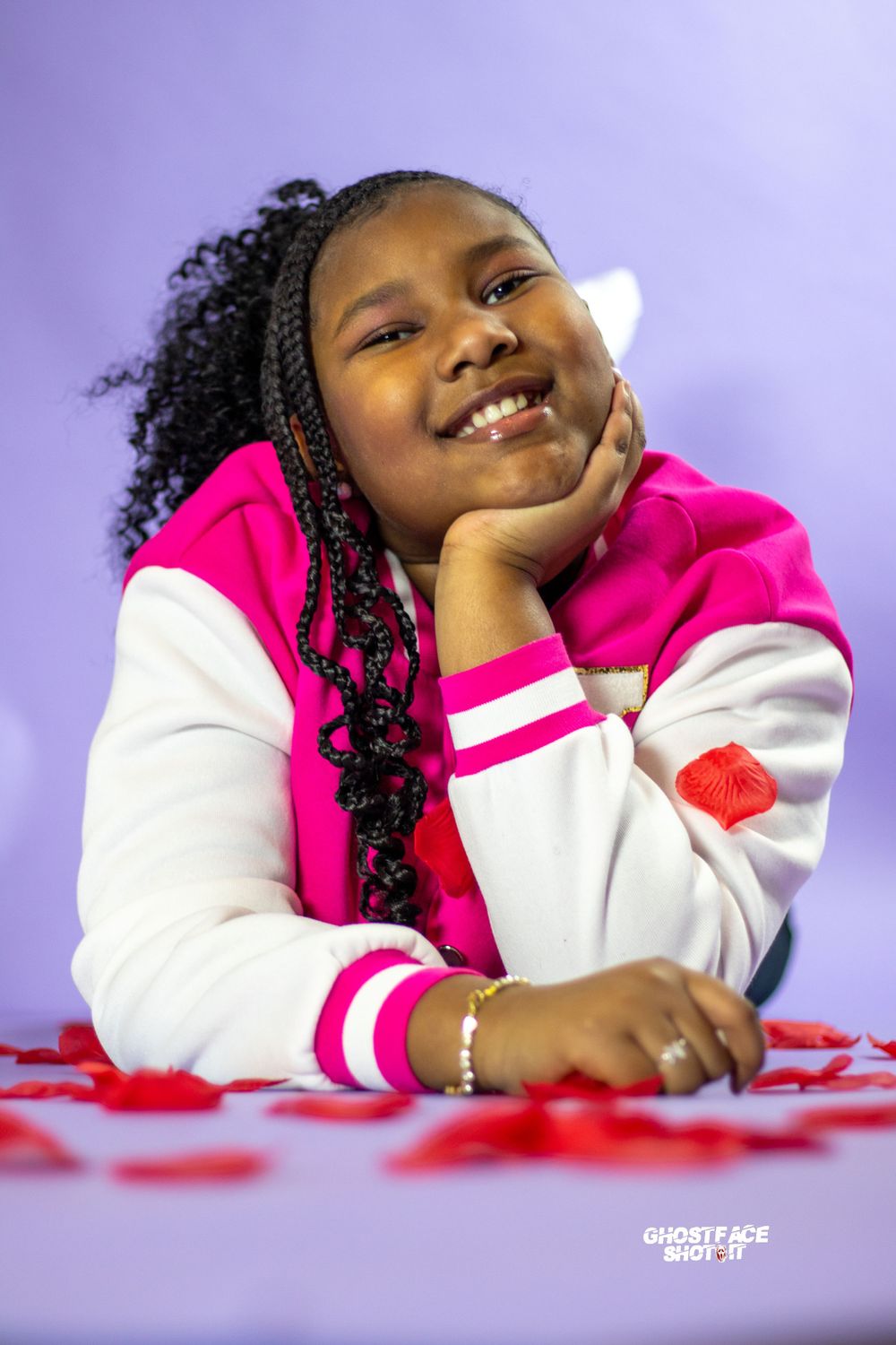 A portrait against a purple background featuring a pink and white varsity jacket and decorative red elements.