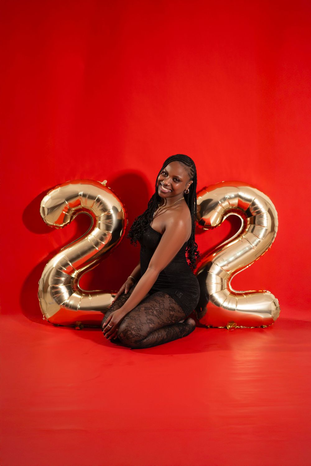 Person in black outfit posing with large metallic number 22 balloons against red background.