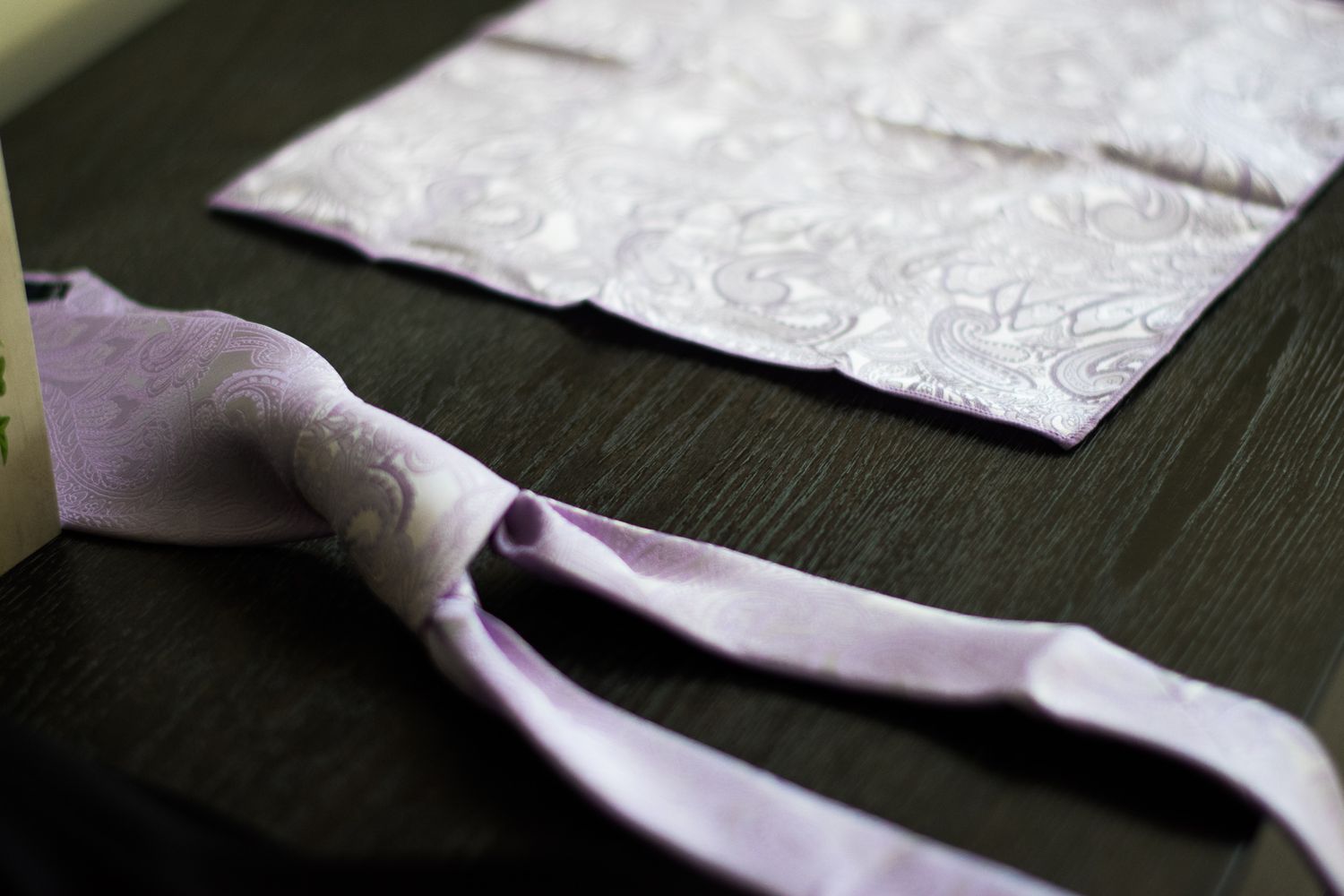 A pale purple tie lies on a dark wooden surface next to patterned paper.
