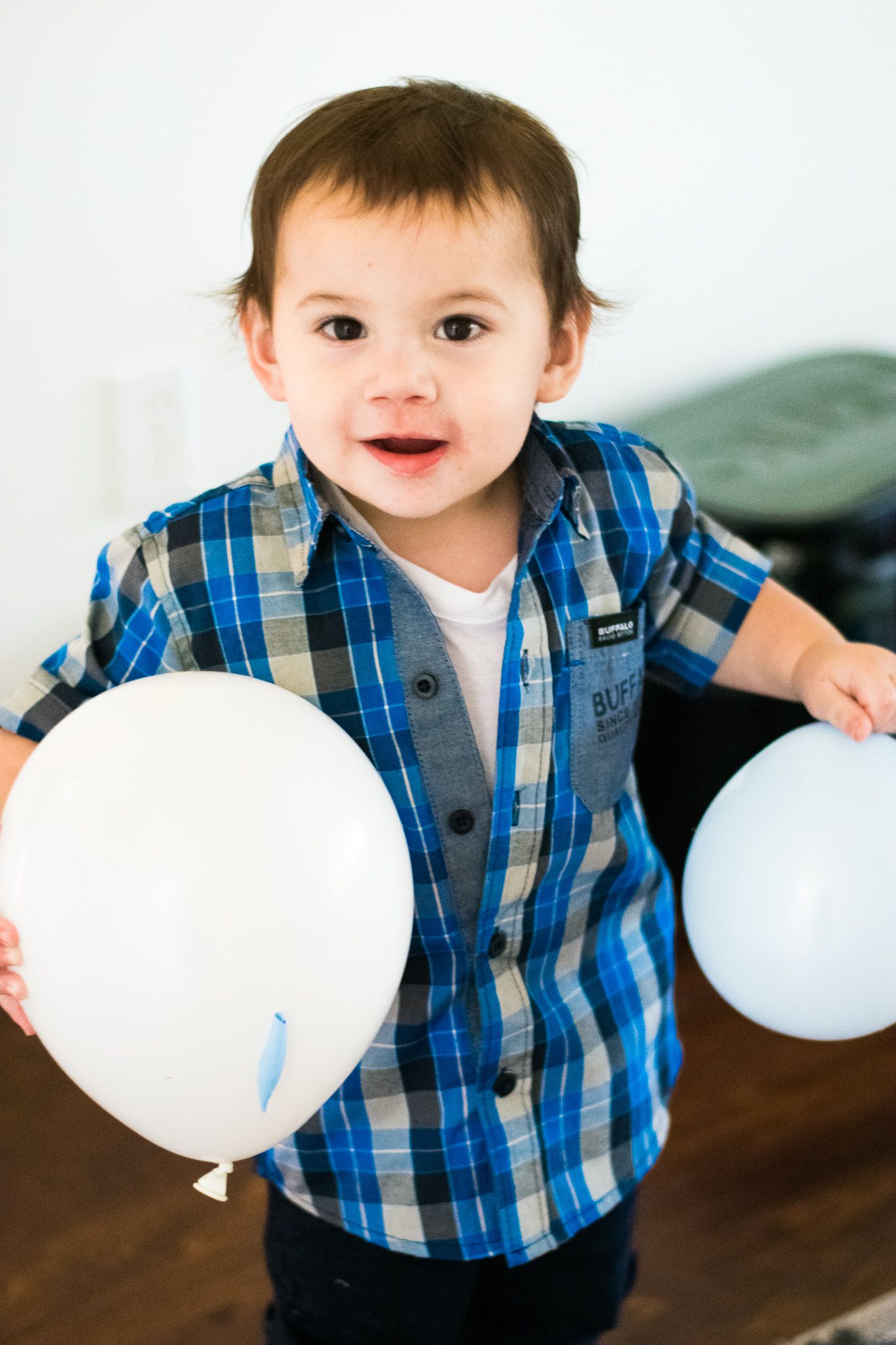 A happy toddler in a blue plaid shirt holds white balloons.