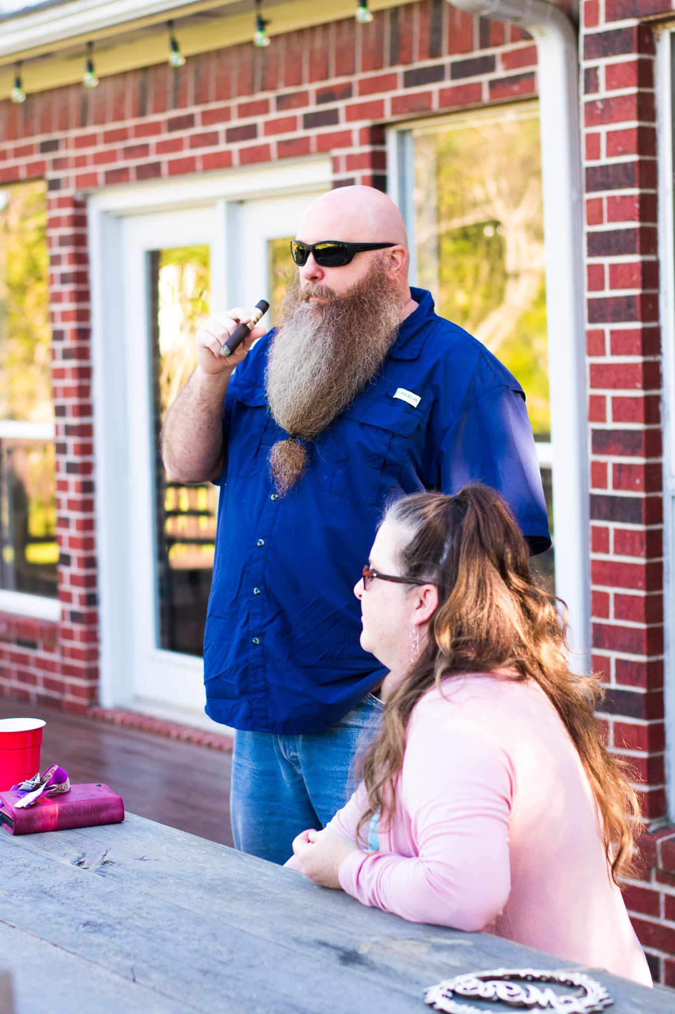 A tall bearded man in blue shirt and sunglasses stands near a brick building.