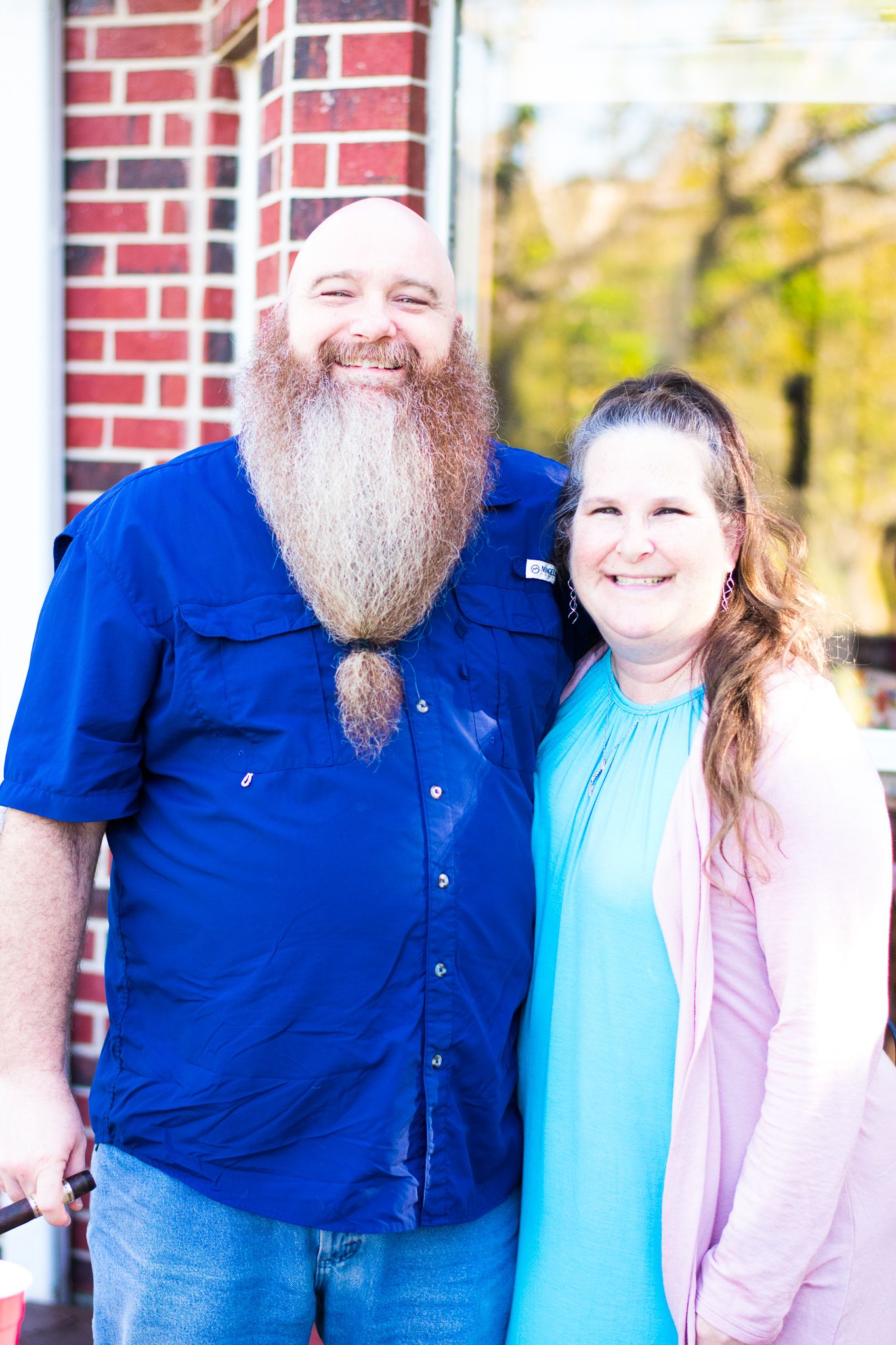 A tall bearded man in blue shirt stands by brick wall on sunny day.