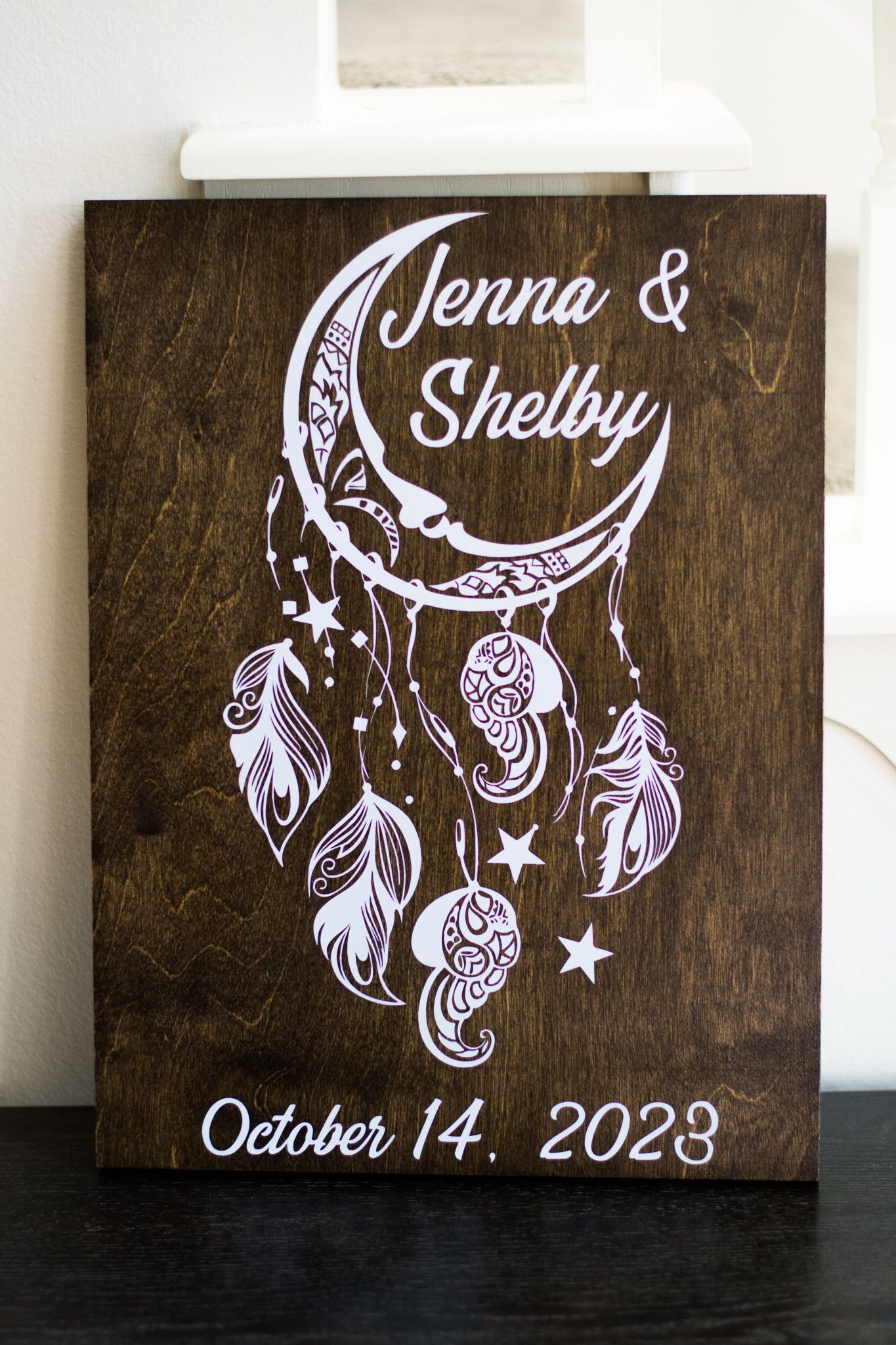 A wooden wedding sign with dreamcatcher design displays names Jenna & Shelby with October 14, 2023 date.