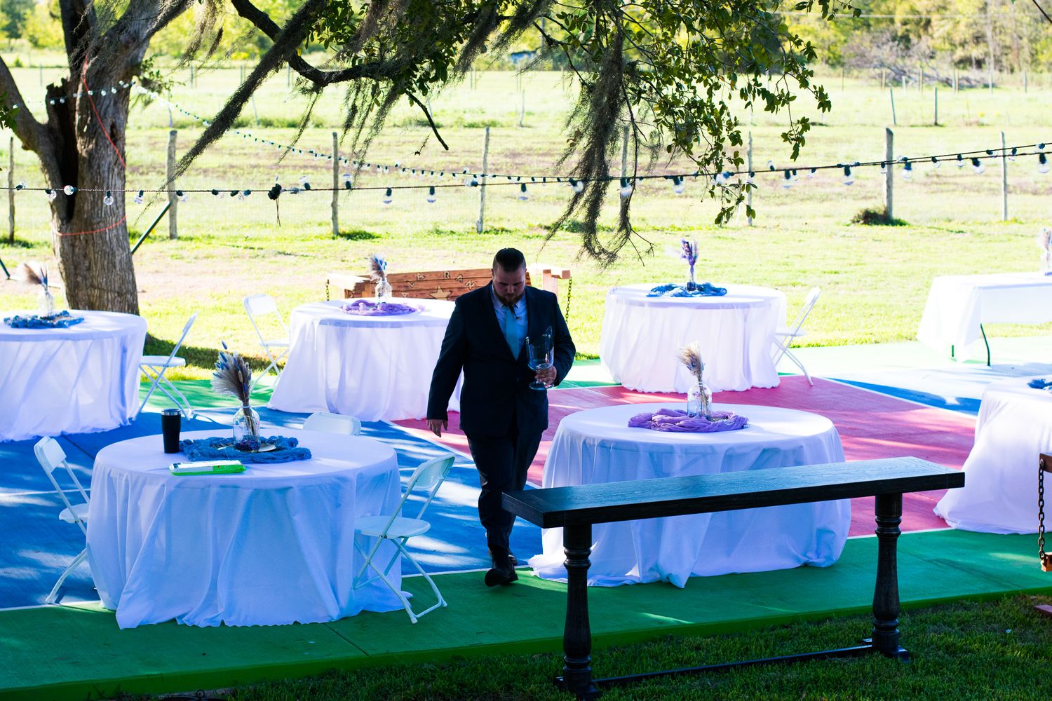 Outdoor event setup with white tables and chairs under trees.
