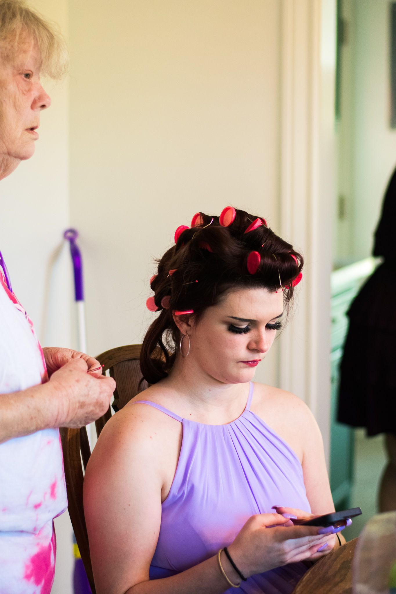 A woman in a purple dress getting her hair styled with red rollers while checking her phone.