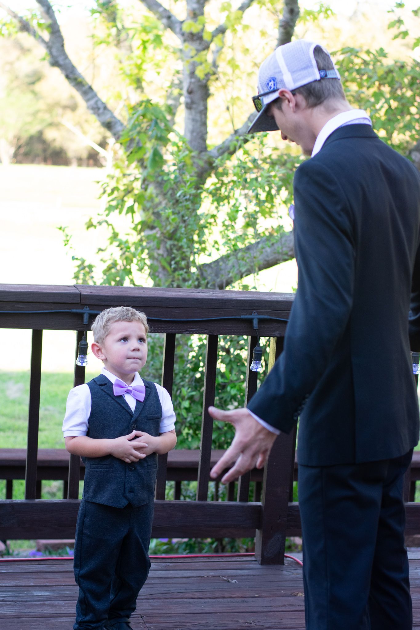 A formal outdoor interaction between an adult in a suit and baseball cap and a young boy in a vest.