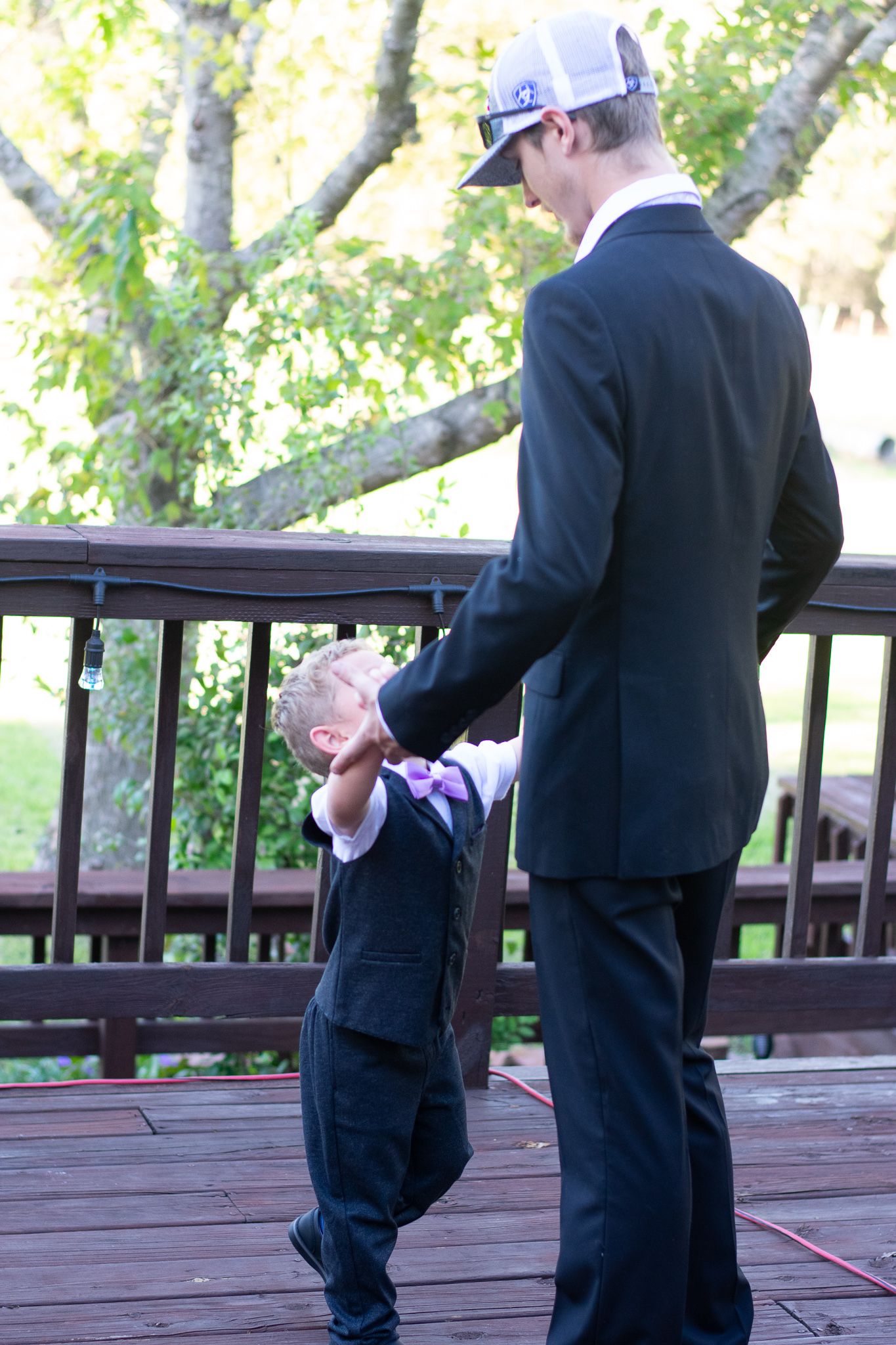 Two figures in formal wear sharing a moment on a wooden deck with trees in the background.