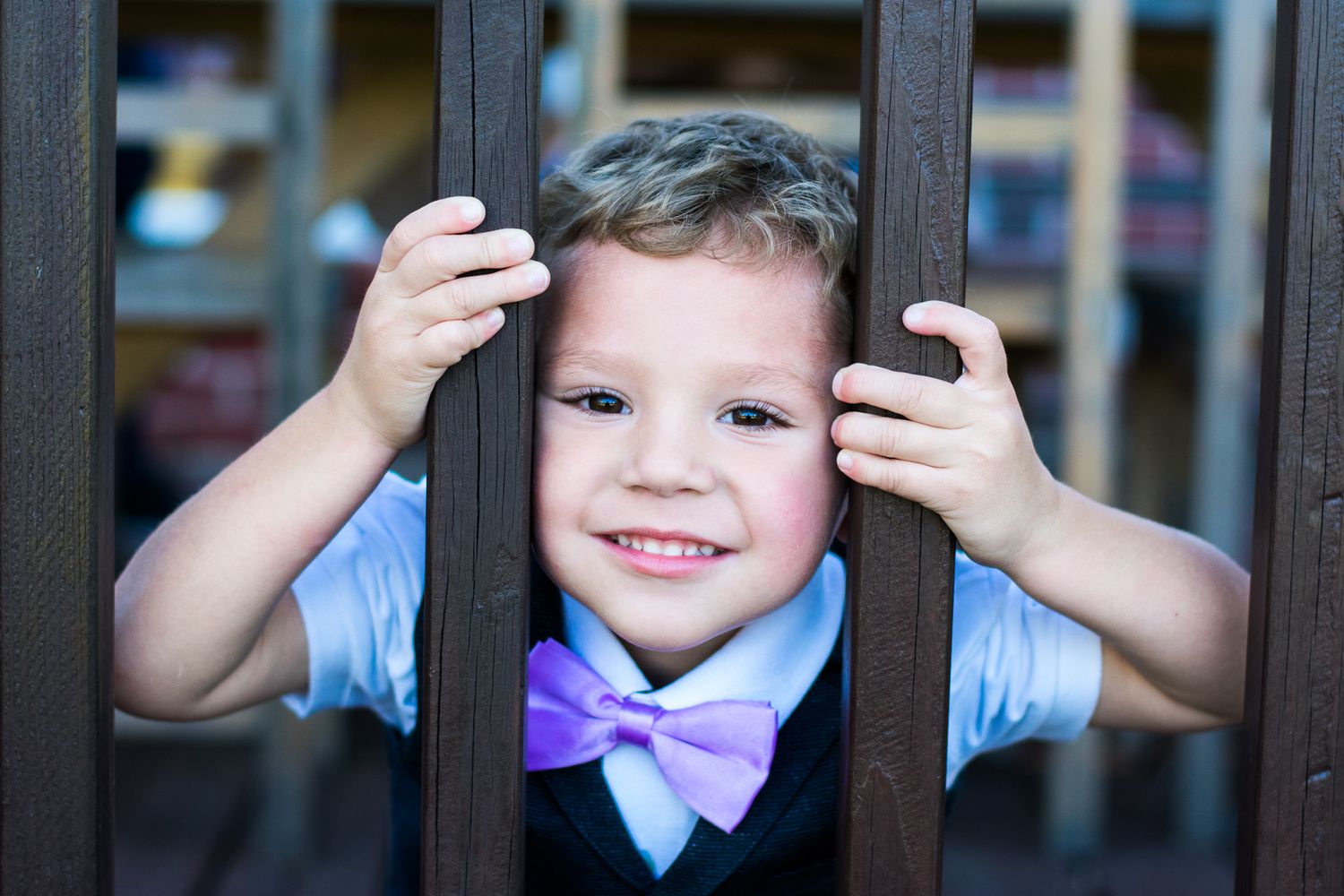 A young child in a purple bowtie peeking through dark railings with a bright smile.