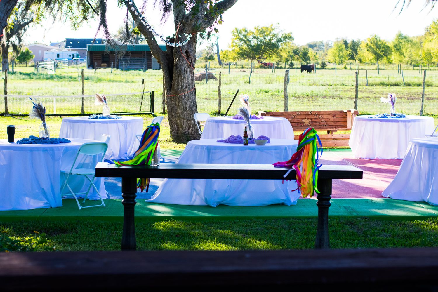Outdoor reception setup with white tablecloths and colorful chairs under trees.