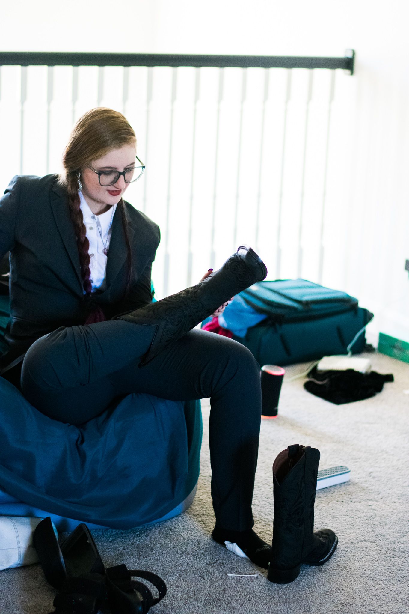 Person in business attire sitting with luggage and boots on floor.