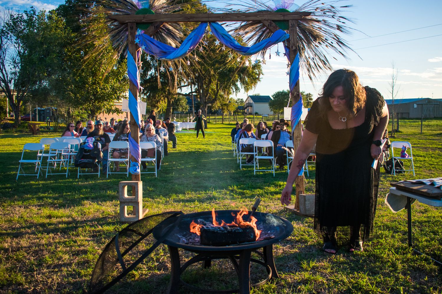 Person tends to outdoor fire pit beneath decorated wedding arch at sunset.