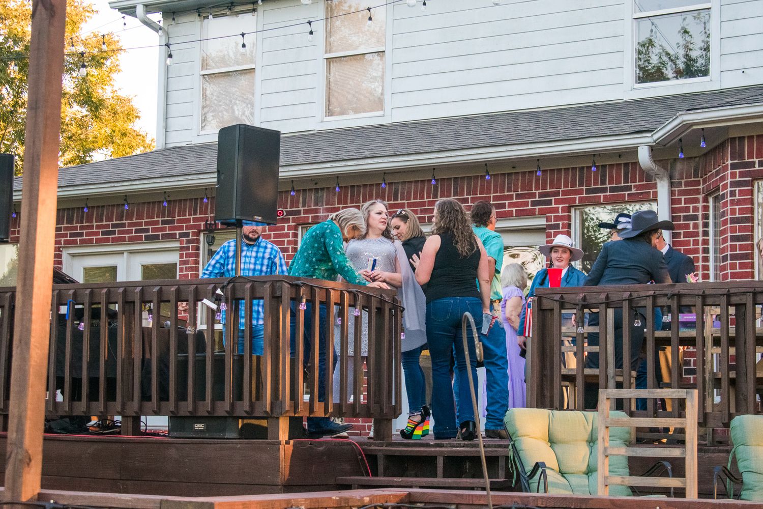 Group gathered on a wooden deck of a brick house during an evening event.