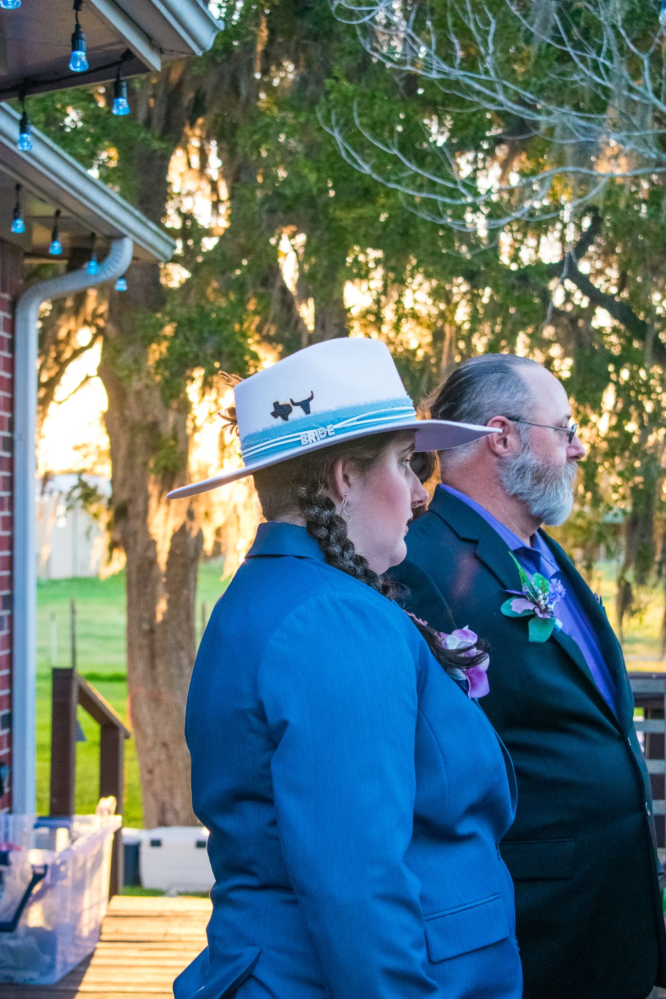 Two people in formal attire standing at sunset with string lights visible.