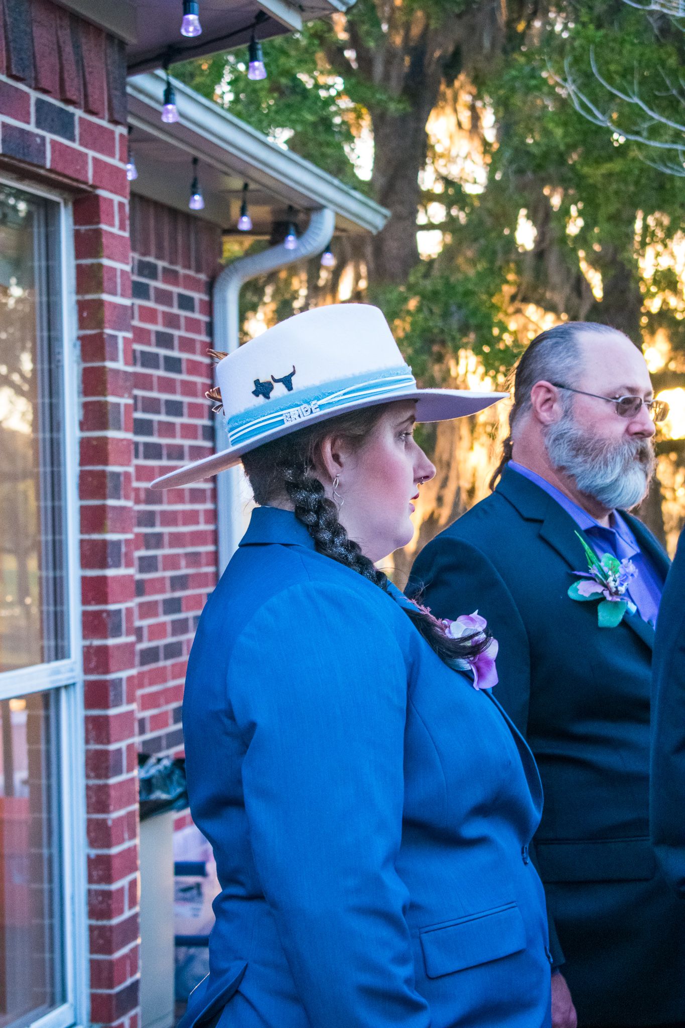 Side view of two people in formal wear standing against brick building with string lights.