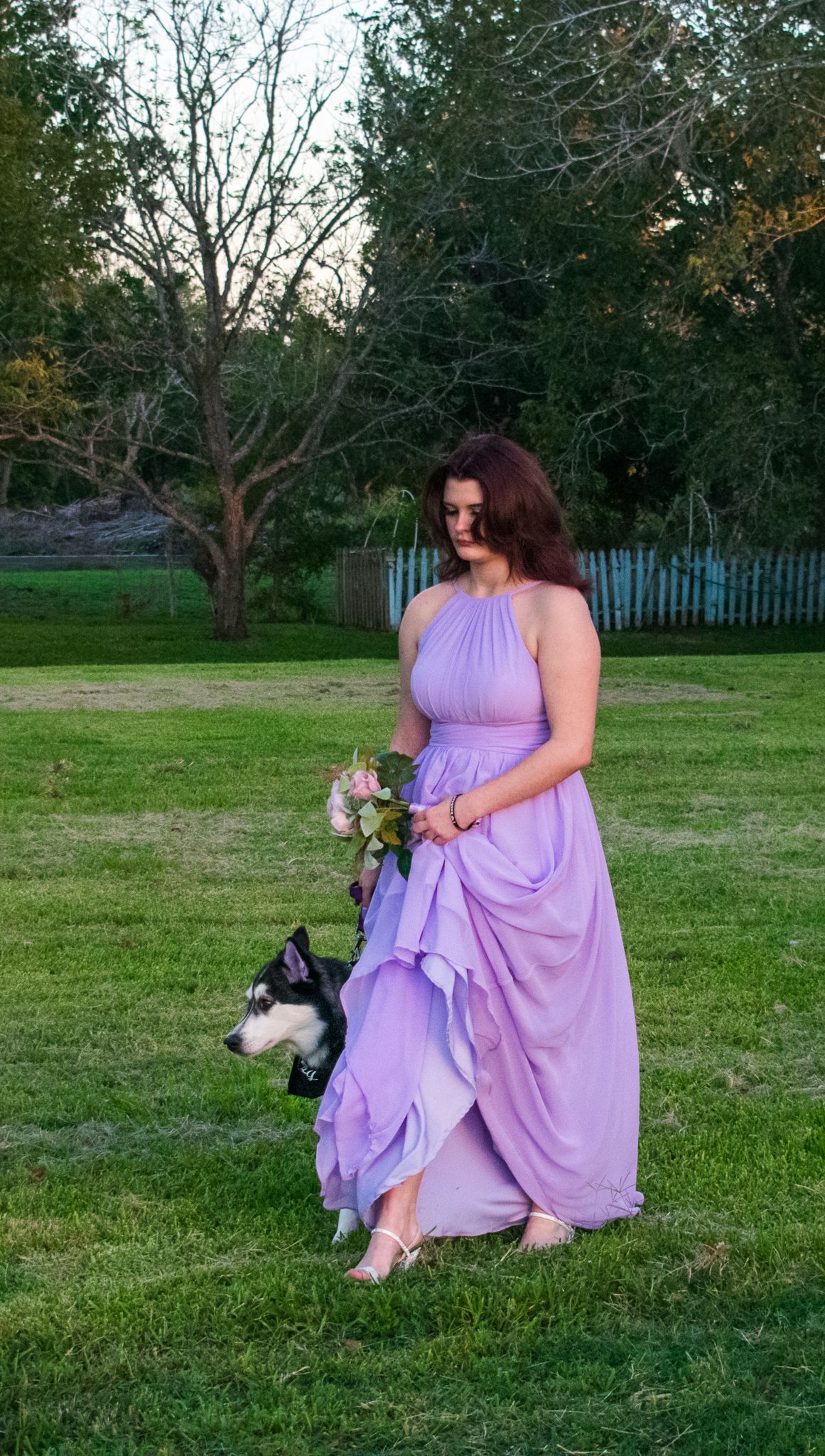 A guest in a flowing lavender gown walks with a small dog on a grassy lawn at dusk.