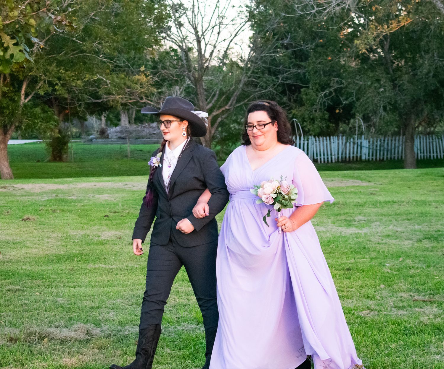 A couple walks arm in arm on a lawn, one in formal black attire and the other in a lavender dress.