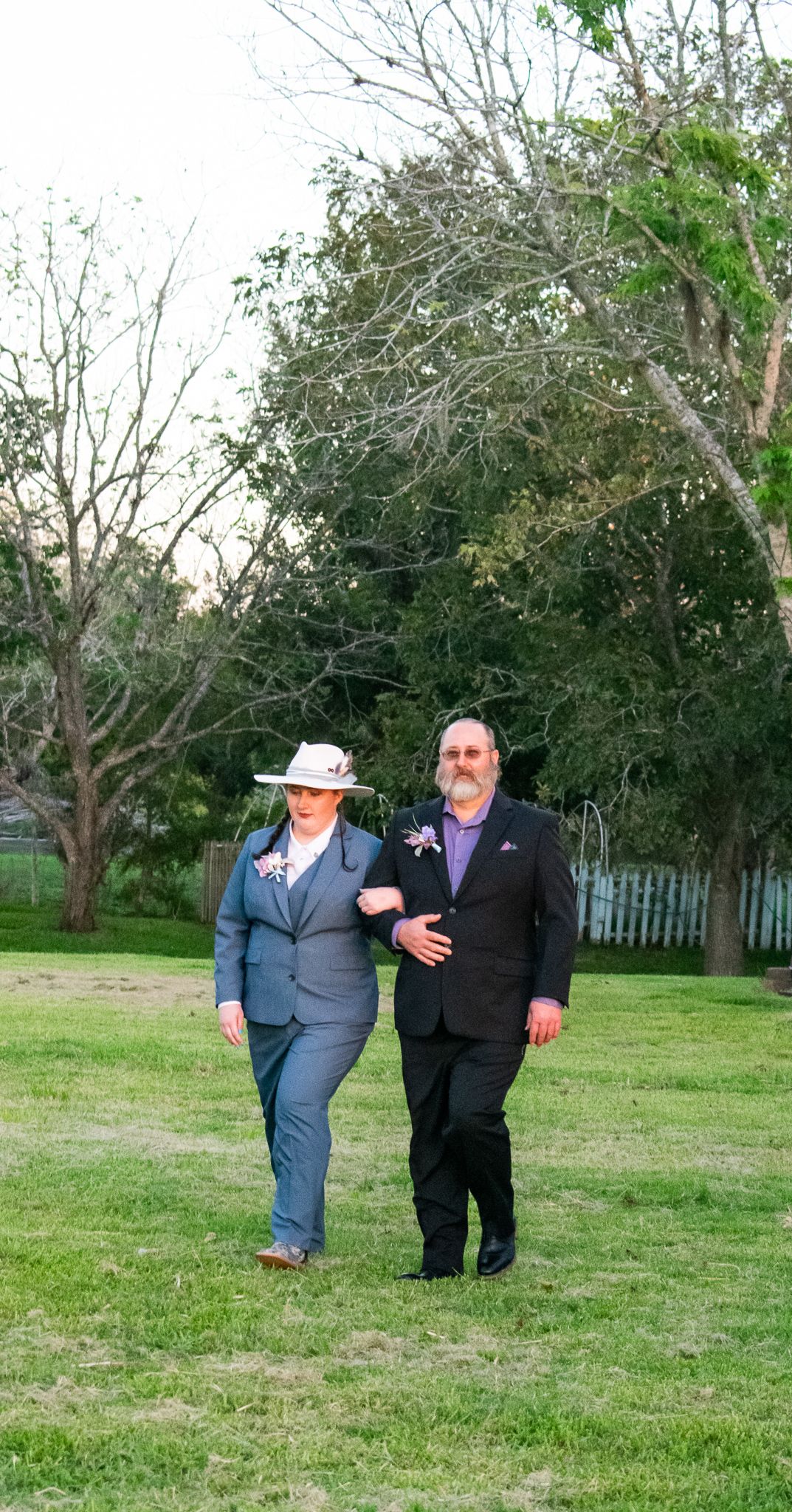 A couple walking arm in arm through a garden setting wearing formal attire.