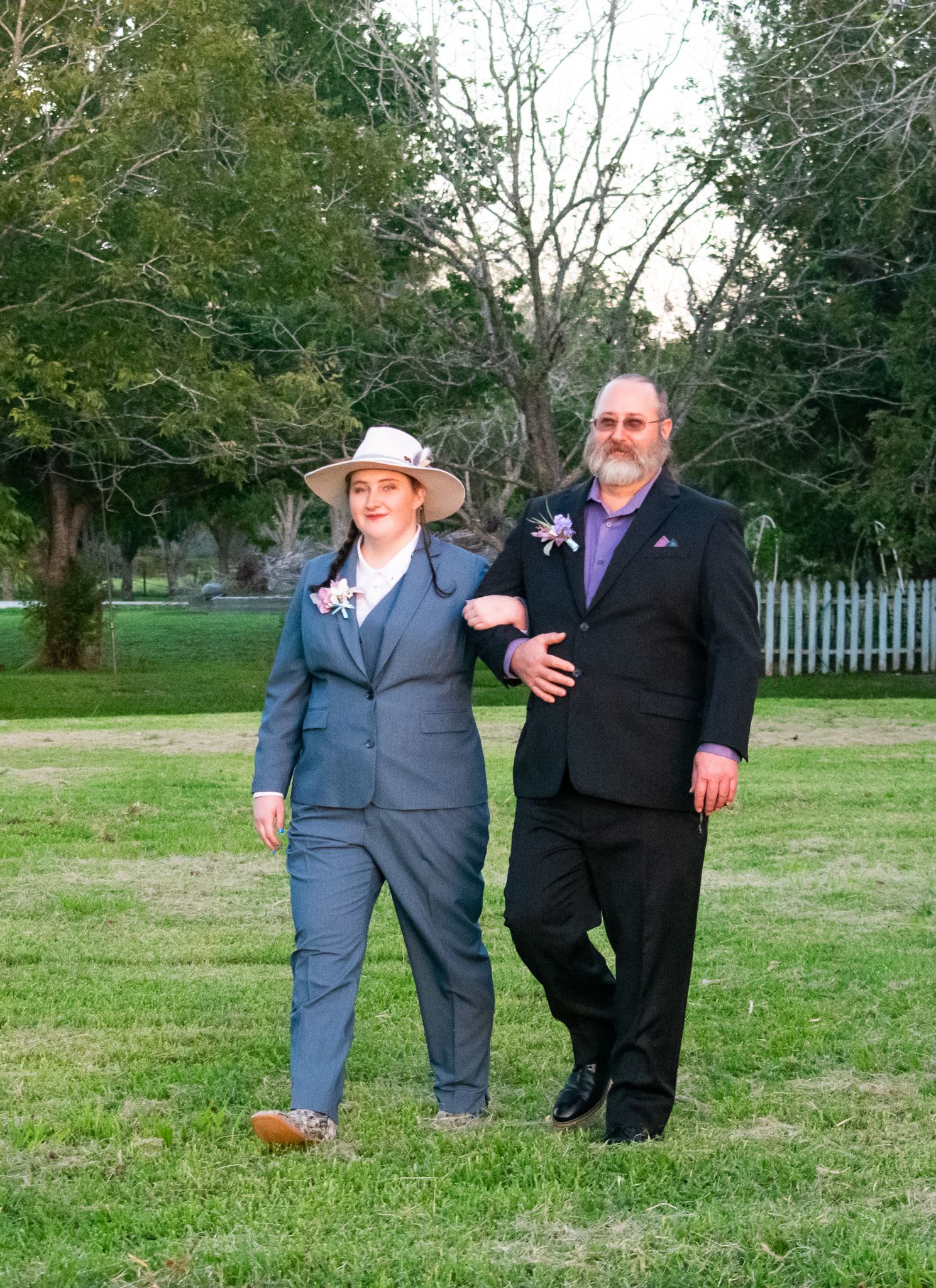 A couple in coordinated formal wear poses together on a grassy lawn with trees in background.
