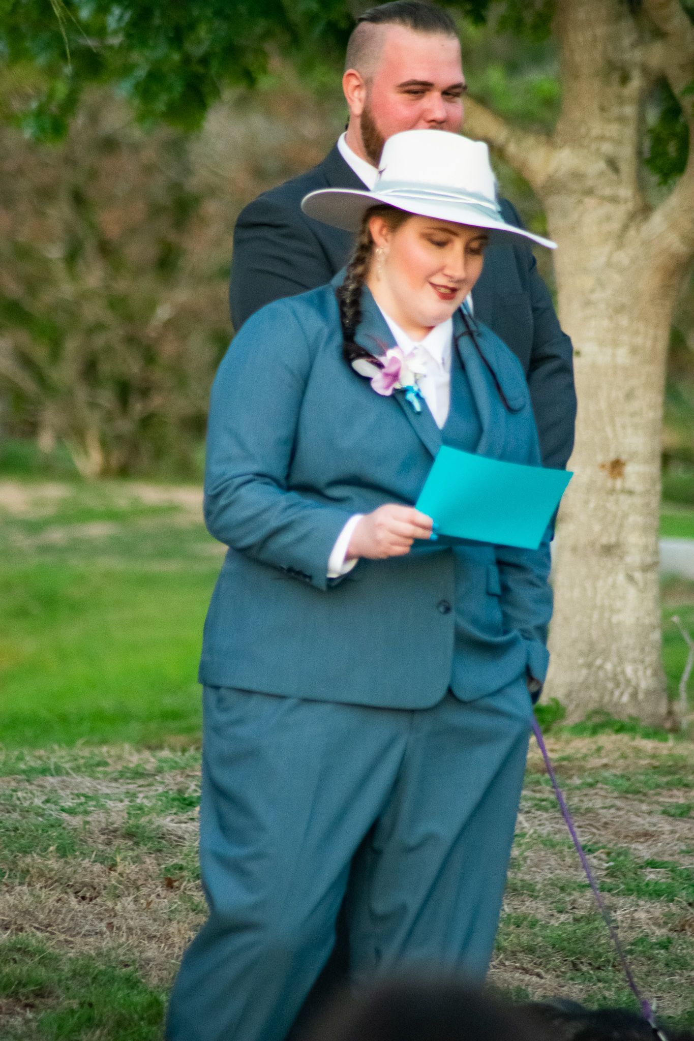 A participant in a teal suit and white hat reading from a turquoise folder during an outdoor ceremony.