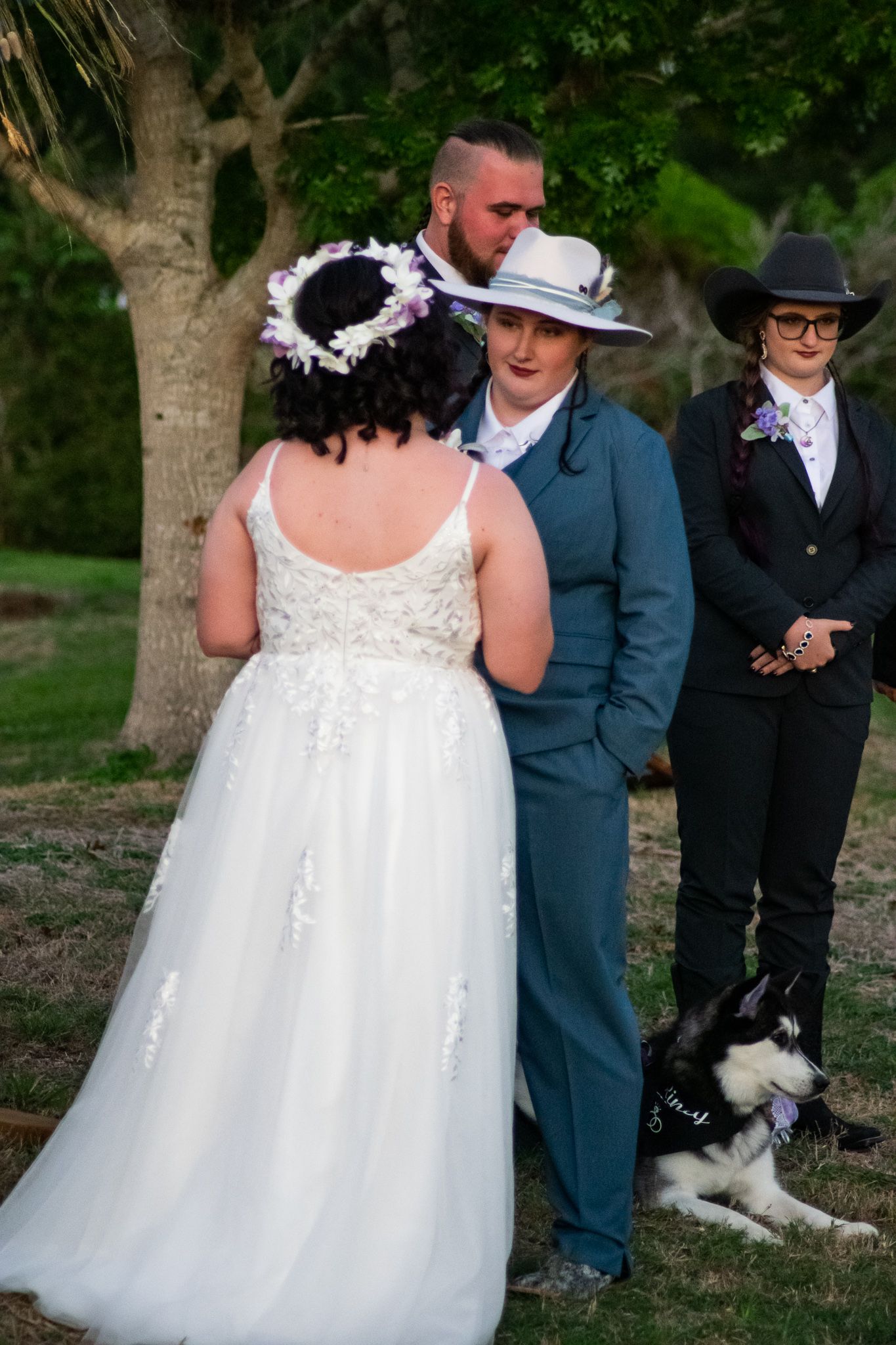 A wedding ceremony with attendees in formal wear and a husky dog present under green trees.