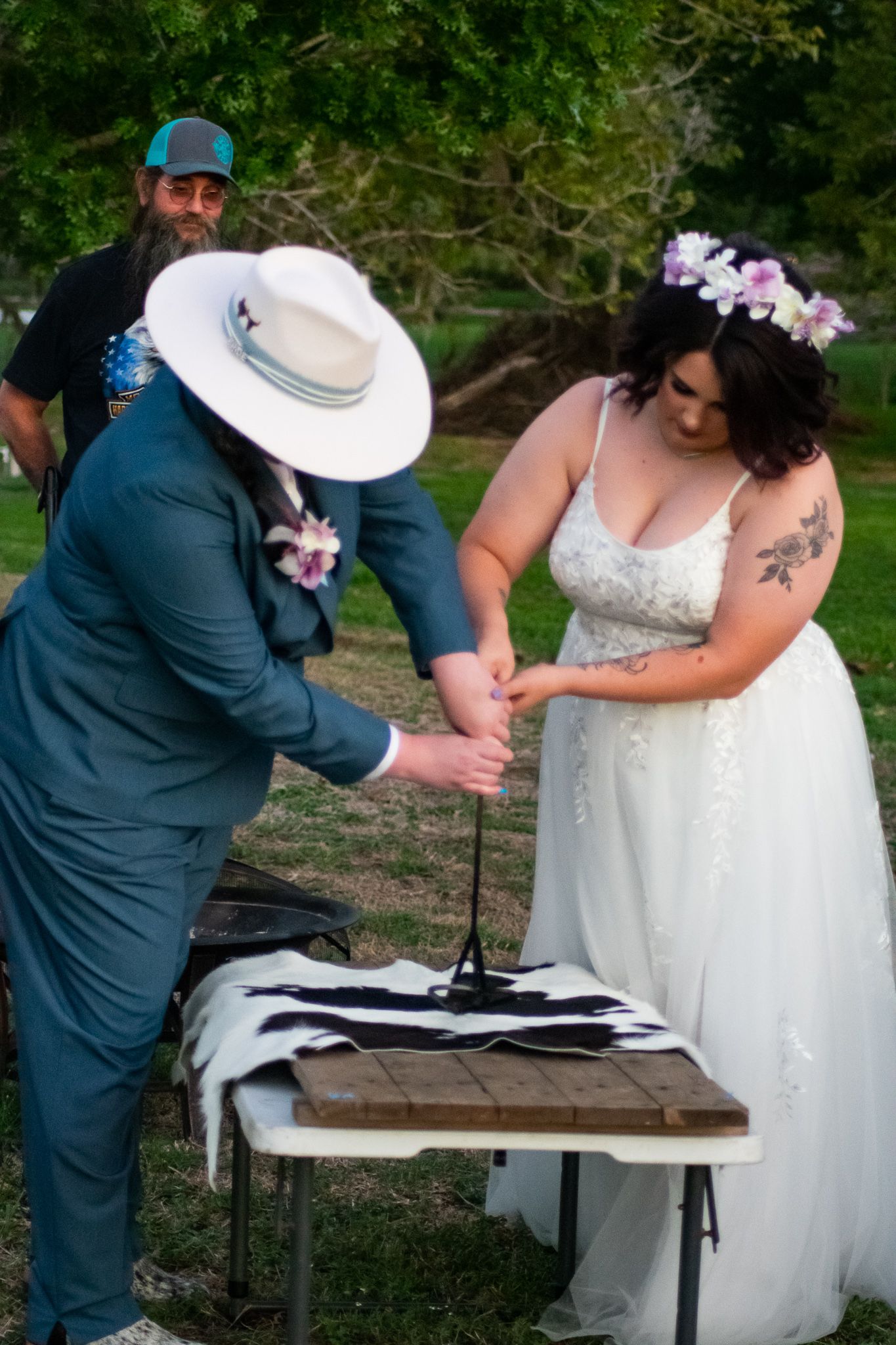 Wedding ceremony participants gather around a wooden table for a unity ritual outdoors.