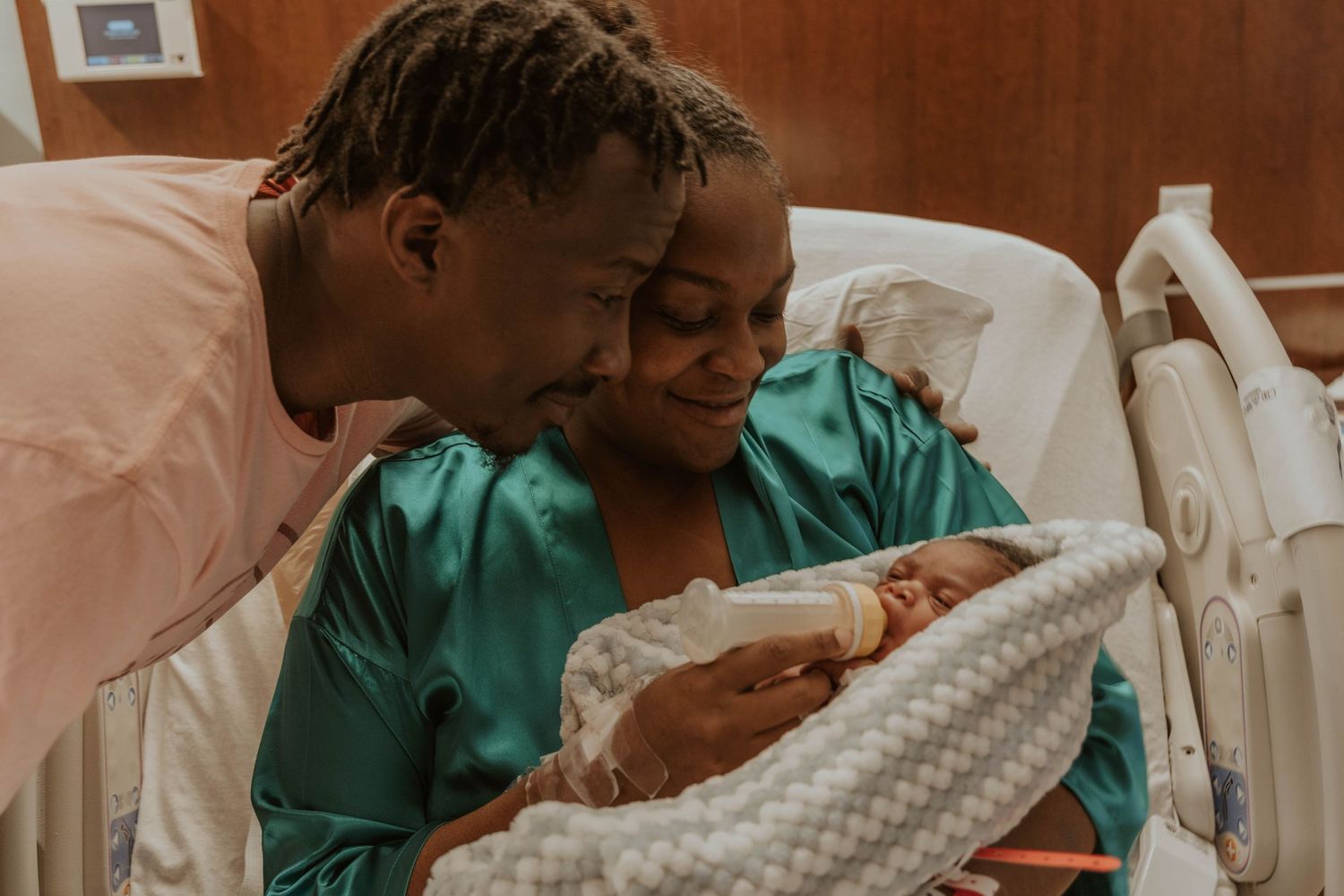 A tender moment in a hospital room as parents embrace their newborn wrapped in a patterned blanket.