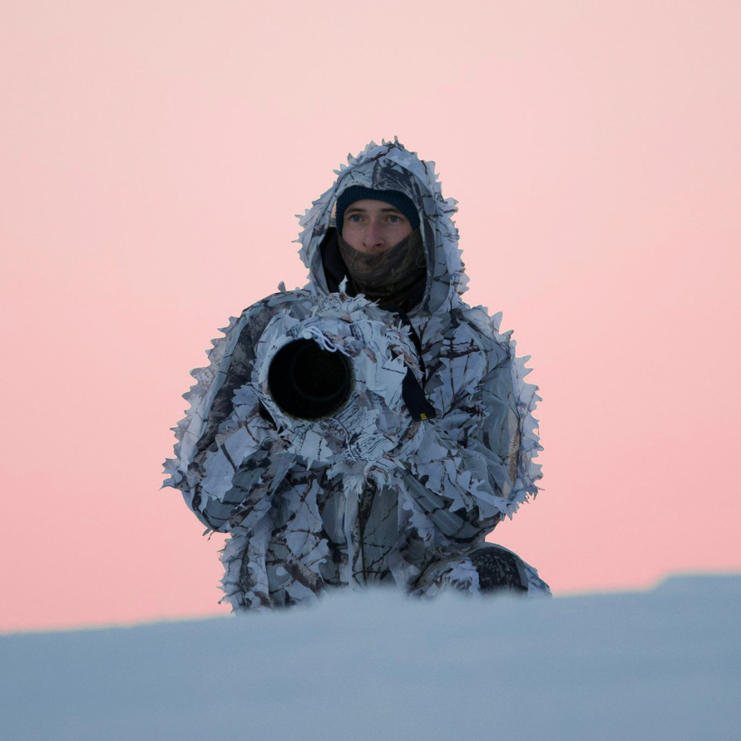 A camouflaged photographer in white winter gear against a pink sunset sky.
