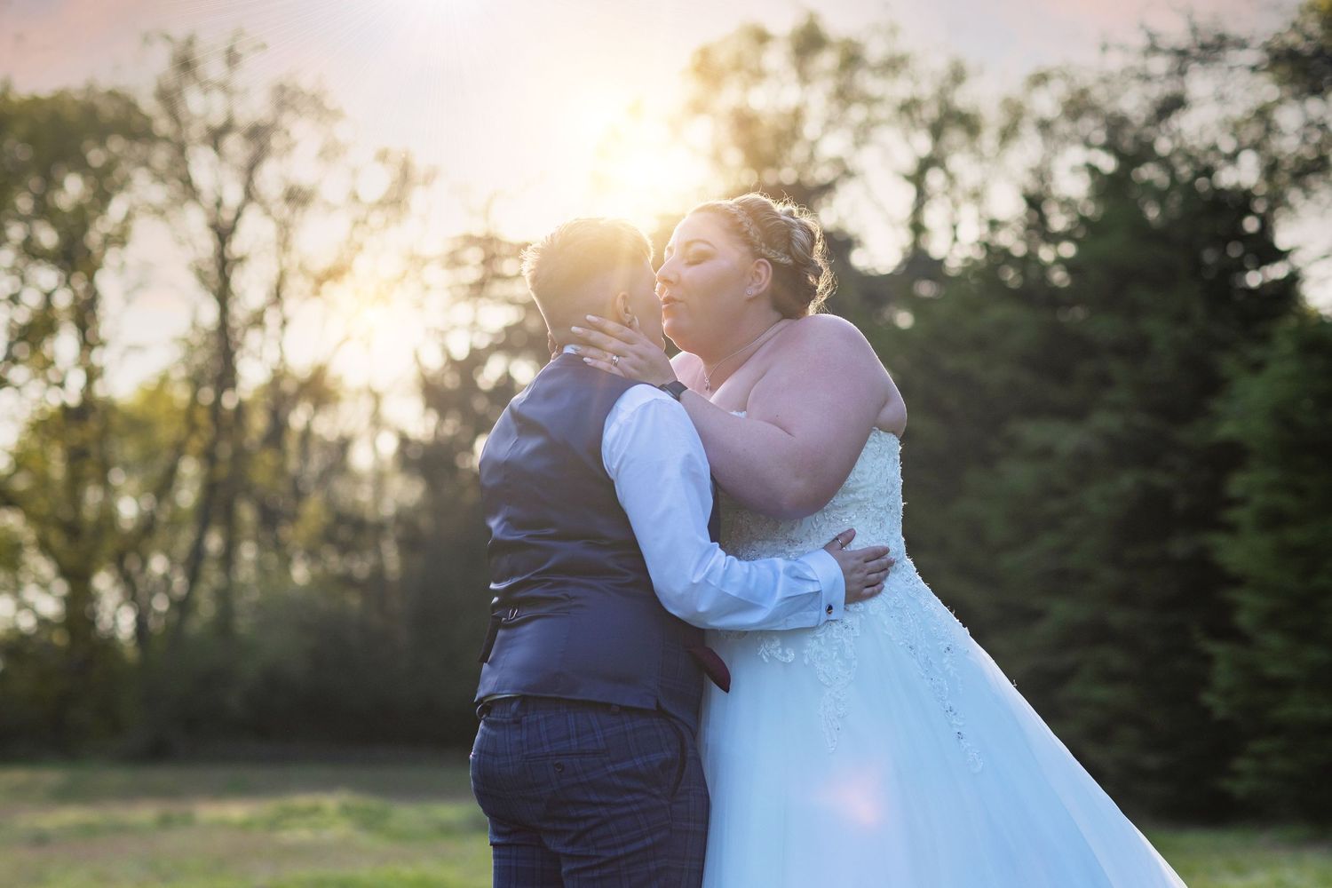 Same-sex couple sharing a kiss outdoors at sunset, with one partner in a white wedding gown and the other in a vest