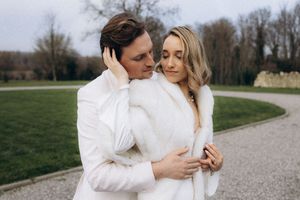 Couple in white outfits embracing outdoors on a winding gravel path through a green landscape.