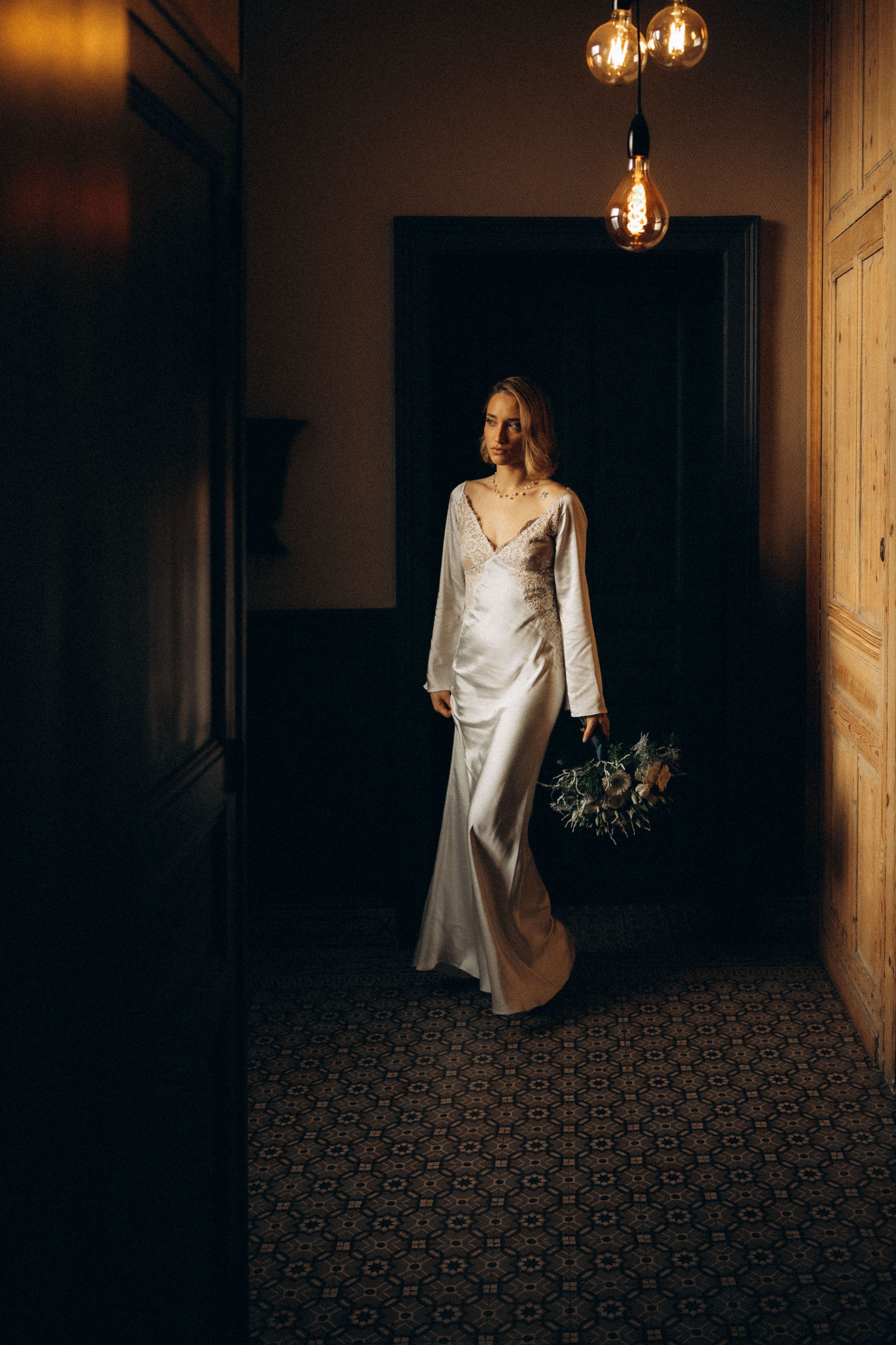 A figure in a white flowing dress walks through a dimly lit hallway with vintage pendant lights and patterned carpet.