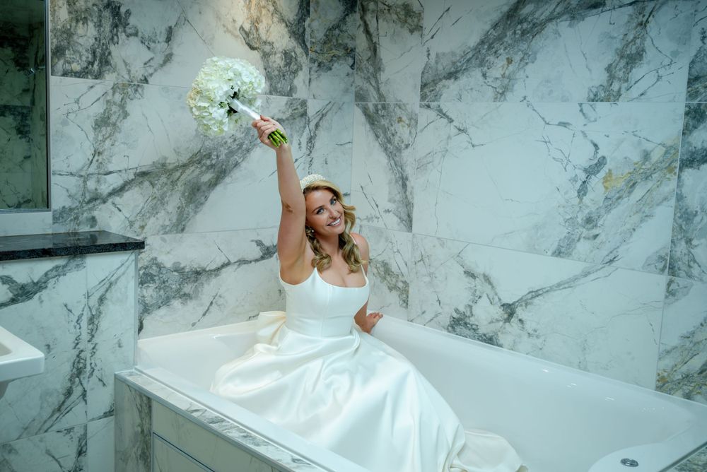 A bride in a white dress raises her bouquet while sitting on a marble surface.