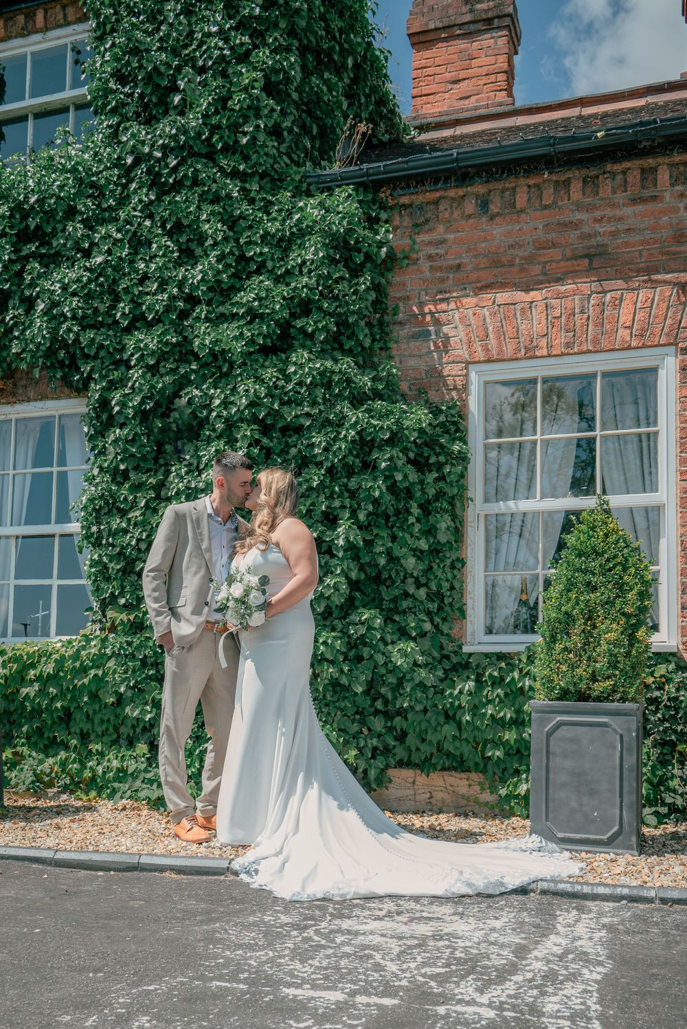 A wedding couple stands in front of a brick building covered in lush green ivy.