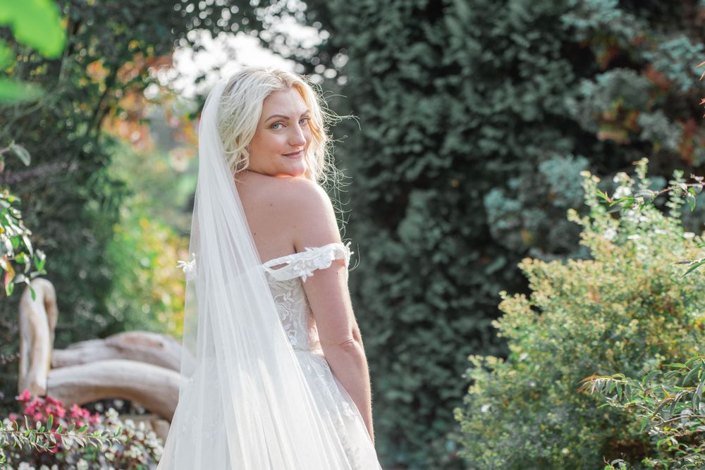 Bride in off-shoulder wedding dress poses against garden greenery backdrop.
