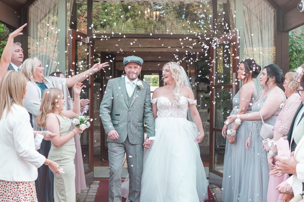 Wedding guests throw confetti as newlyweds exit their ceremony under a covered walkway.