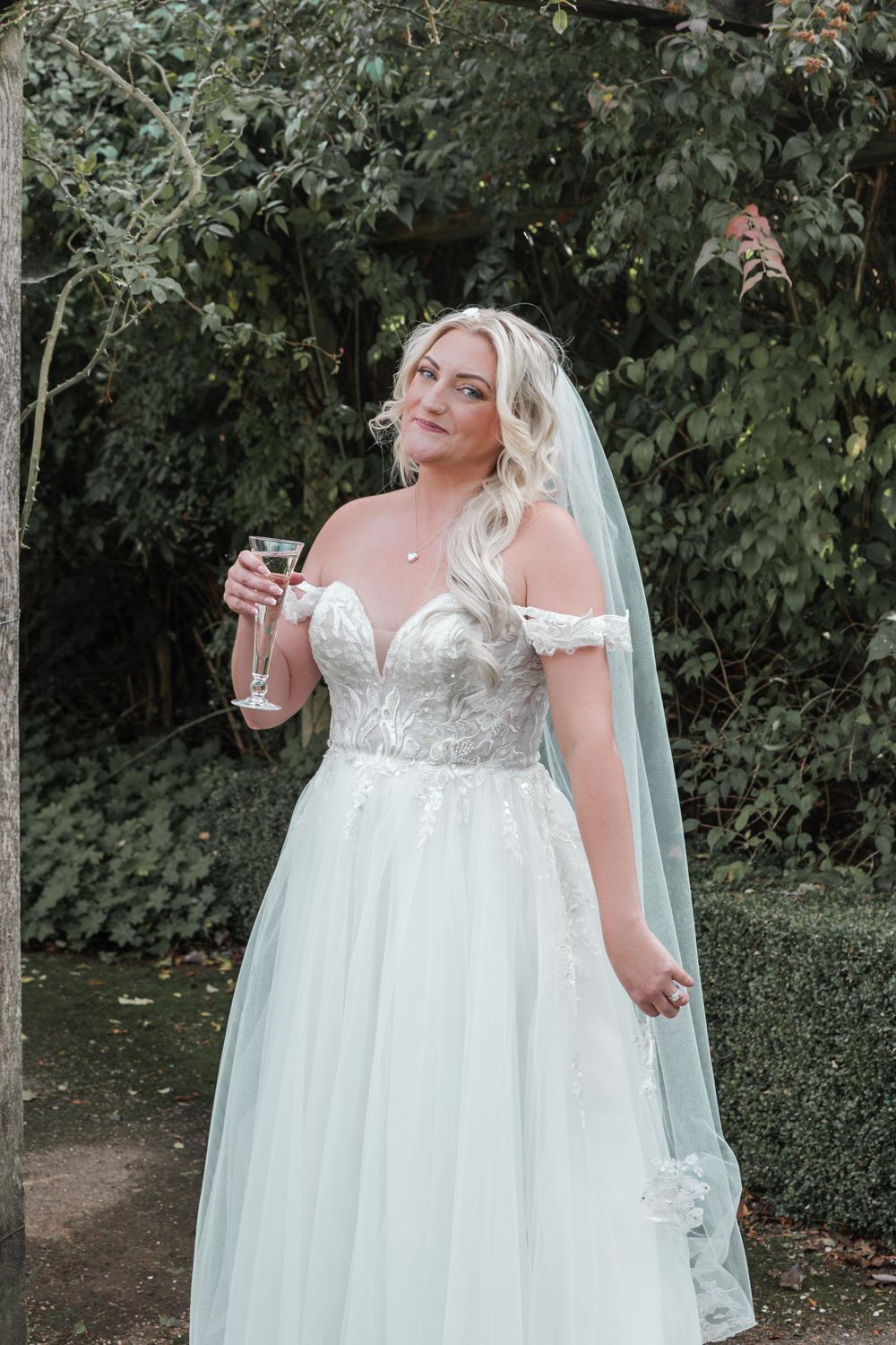 A bride in an off-shoulder lace wedding gown poses with champagne glass in garden setting.