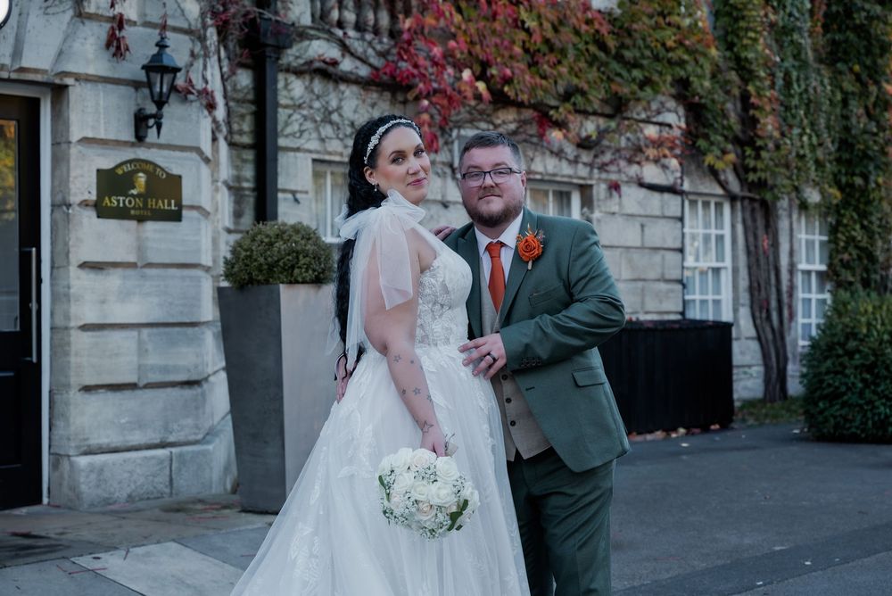 A happy couple poses outside a historic stone building on their wedding day, with fall foliage in the background.