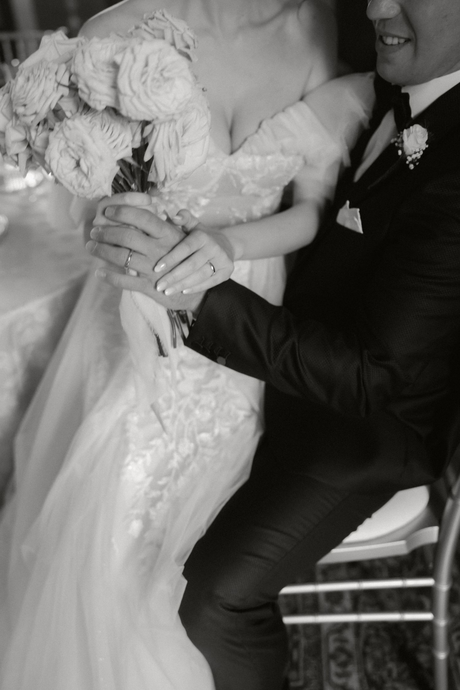 A black and white wedding photo showing hands holding a bouquet of white roses against a lace dress and black tuxedo.