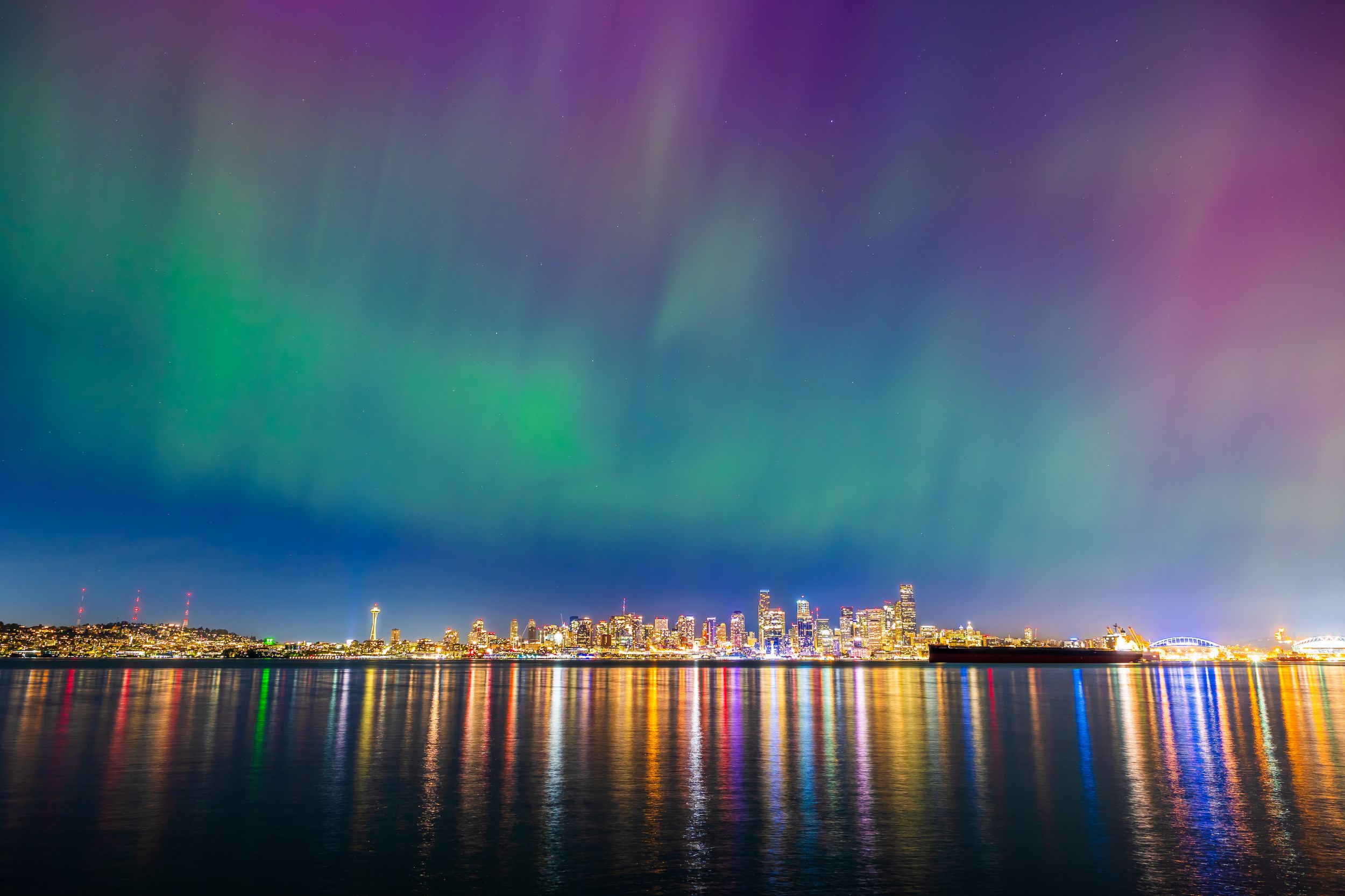 Northern lights dance above a city skyline reflecting colorfully in calm waters at night. Seattle
