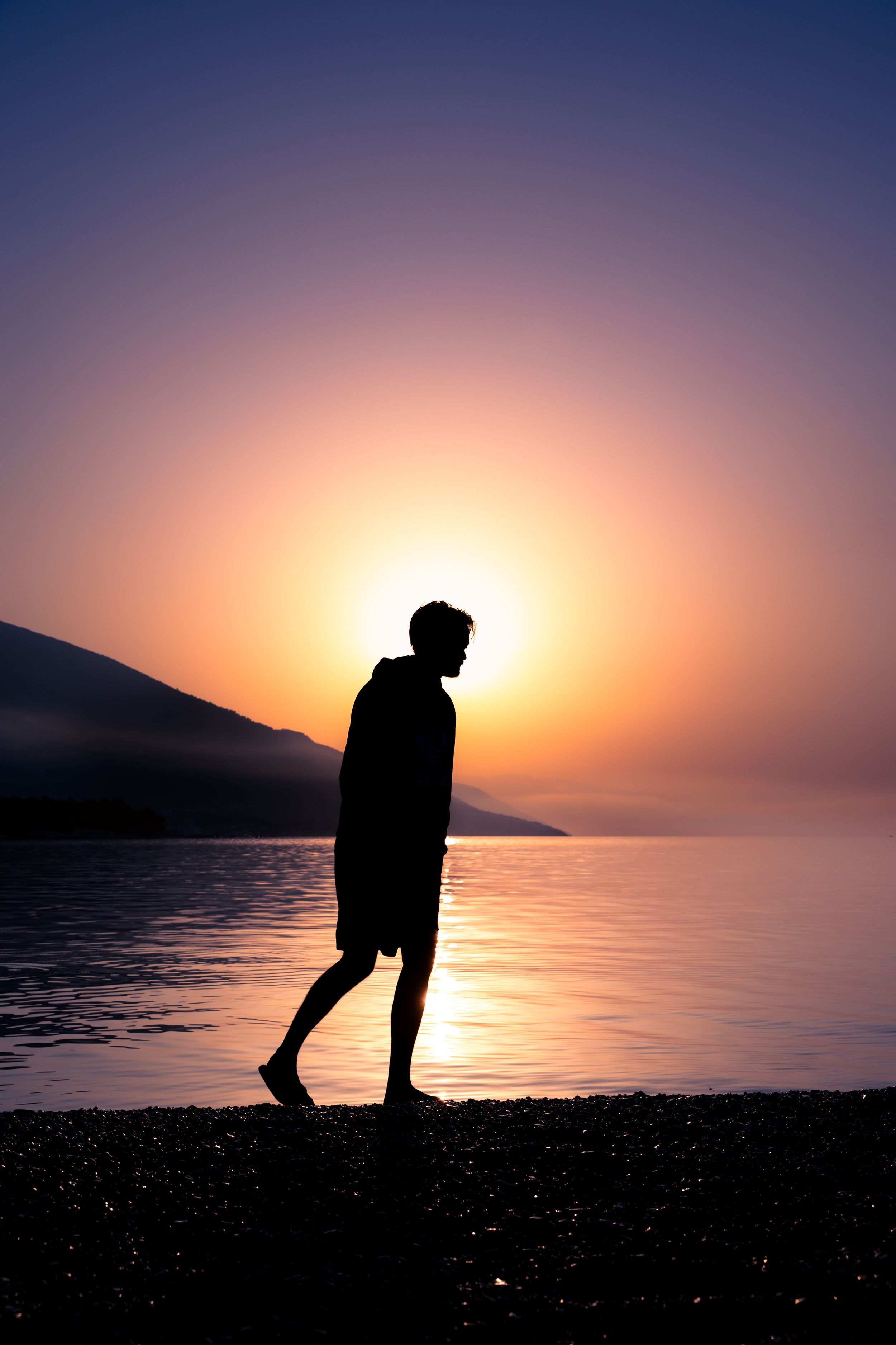 A silhouette of a person walks along the shoreline during a serene sunset over calm waters and mountains.