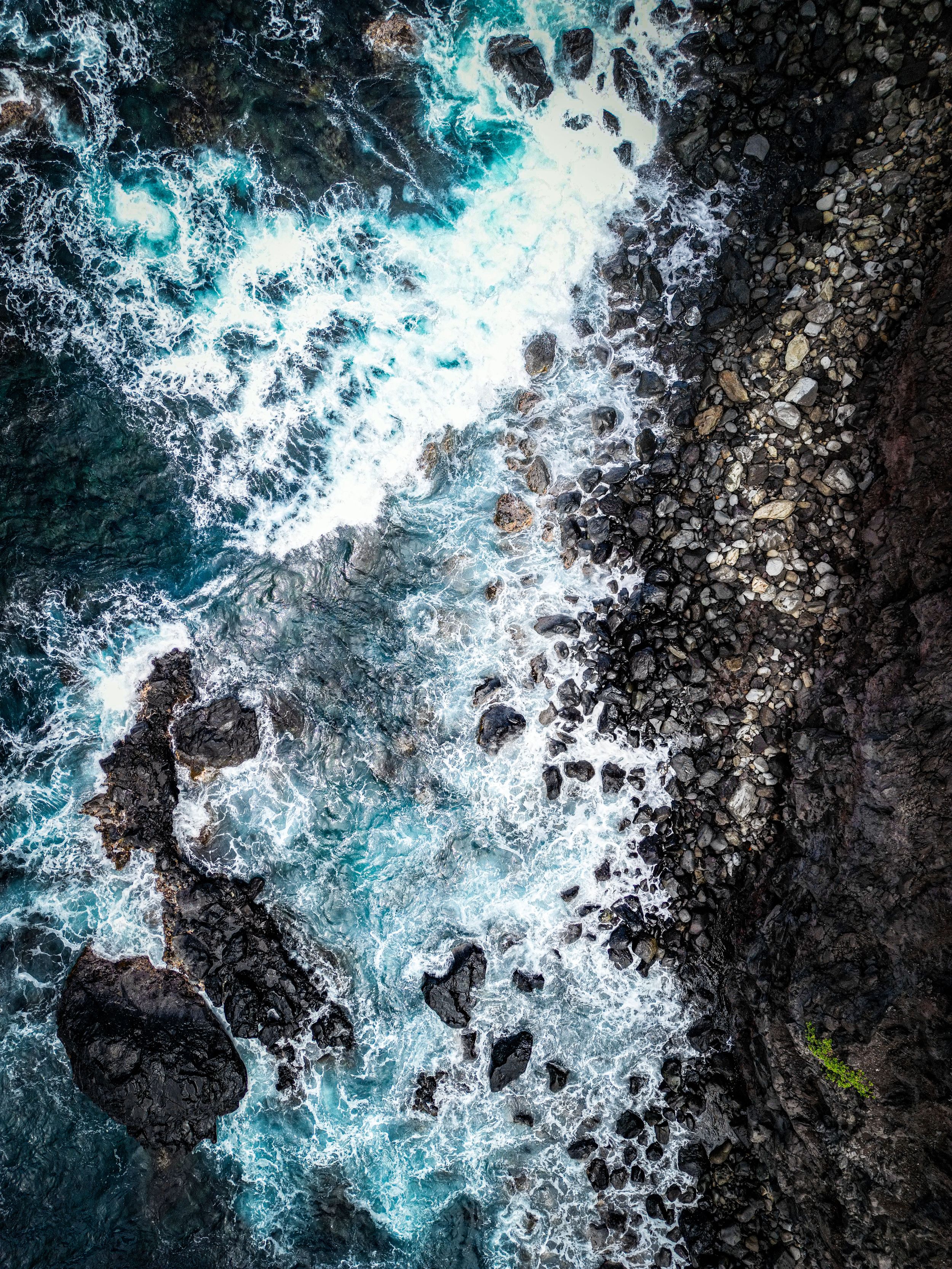Aerial view of turquoise ocean waves crashing against dark volcanic rocks along a dramatic coastline in Maui.