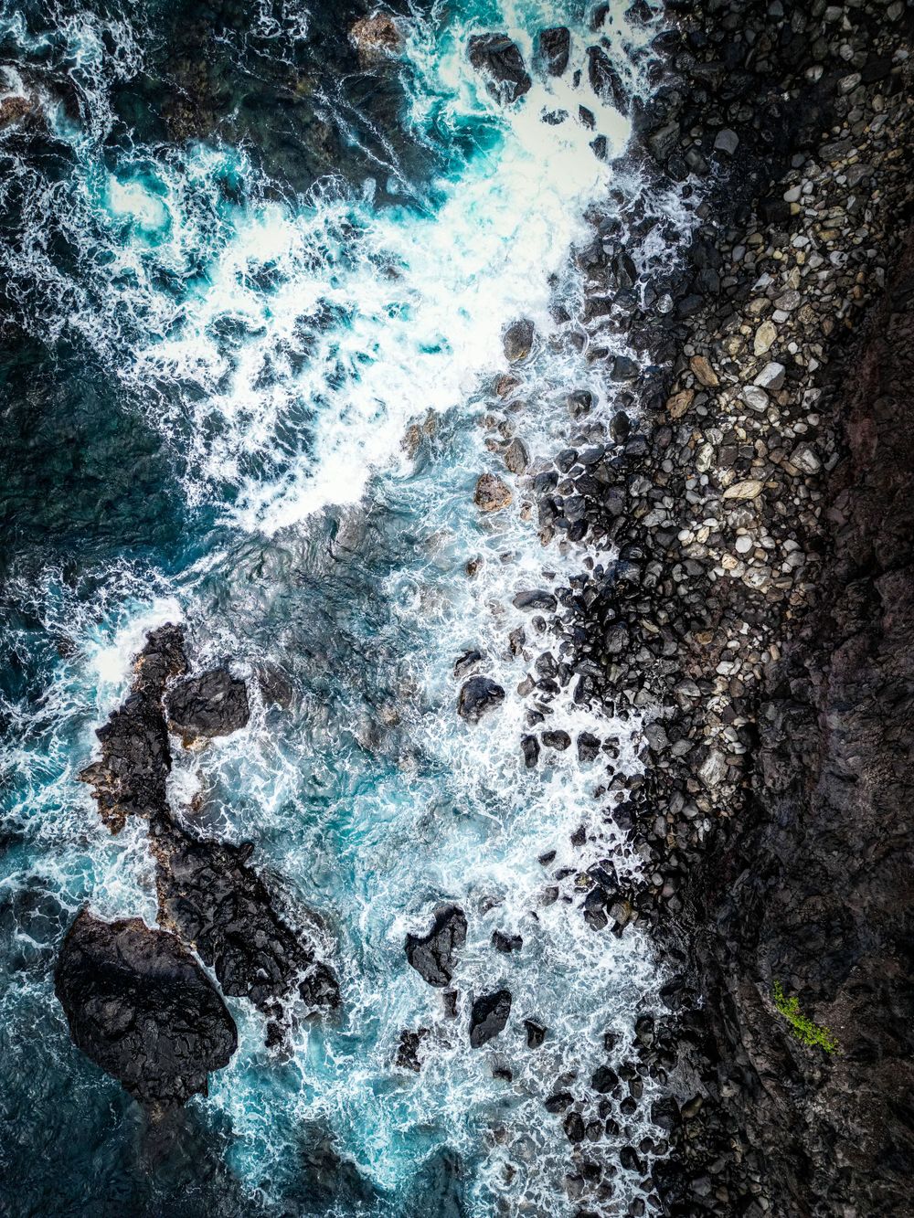 Aerial view of turquoise ocean waves crashing against dark volcanic rocks along a dramatic coastline in Maui.