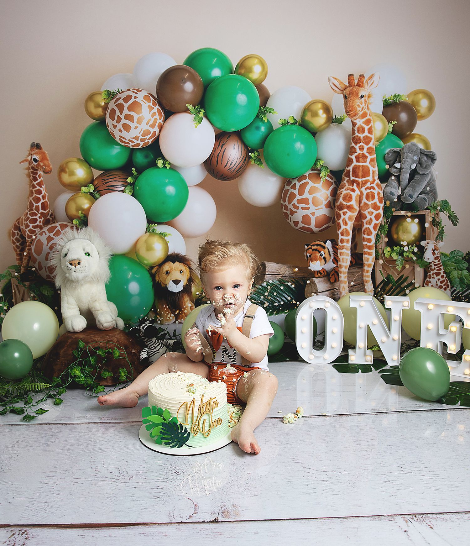 Safari-themed first birthday party setup with green and gold balloon arch, plush animal decorations, and cake smash scene in Bexley