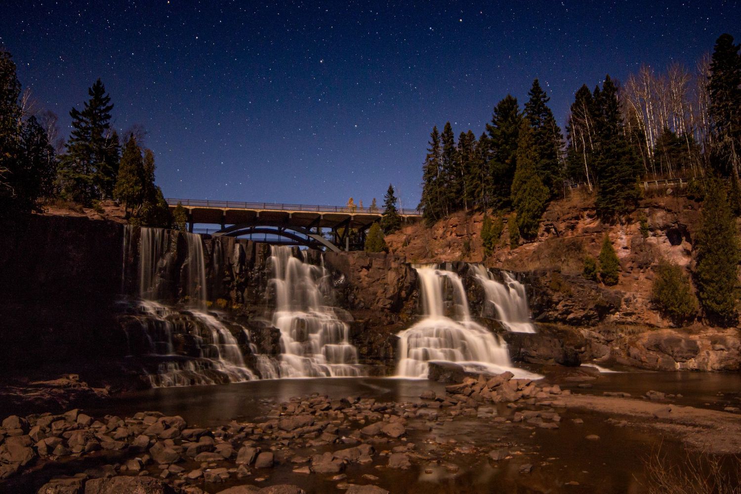 Night sky landscape photography of Gooseberry Falls State Park beneath star-filled sky on a full moon night, dramatic celestial wall art.