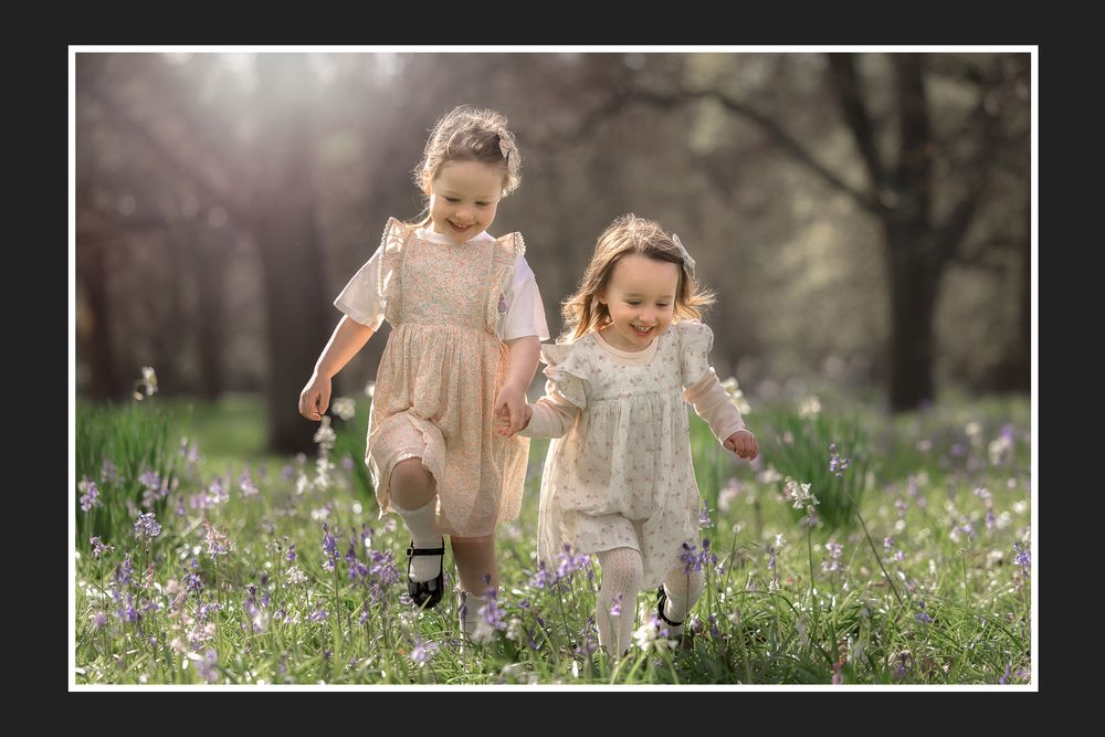 Two young girls in white dresses running through a field of purple wildflowers in soft spring sunlight.