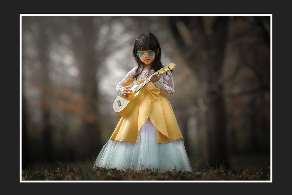 Young girl in golden princess dress plays ukulele outdoors in misty forest setting.